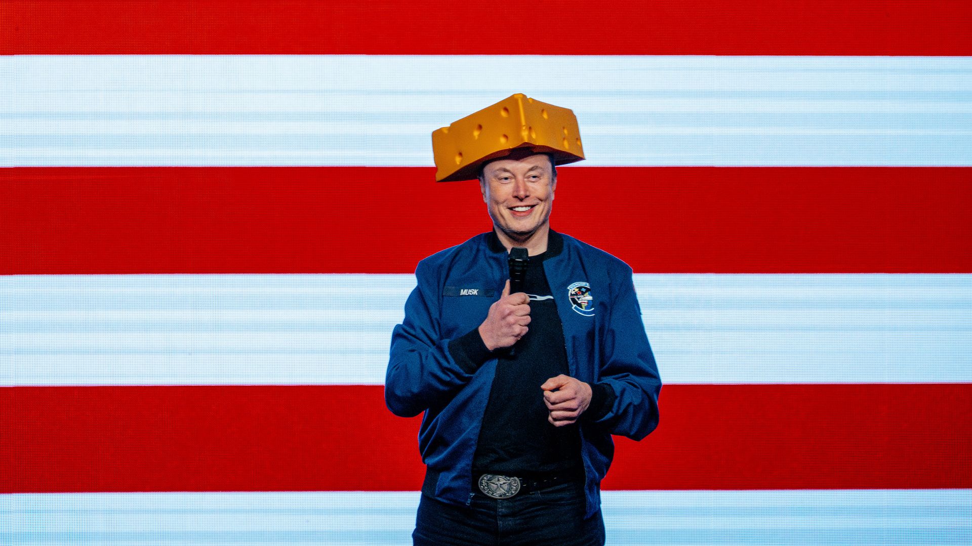 Elon Musk wearing a blue windbreaker and a cheese hat, holding a microphone and standing in front of the red and white stripes of a massive American flag backdrop.
