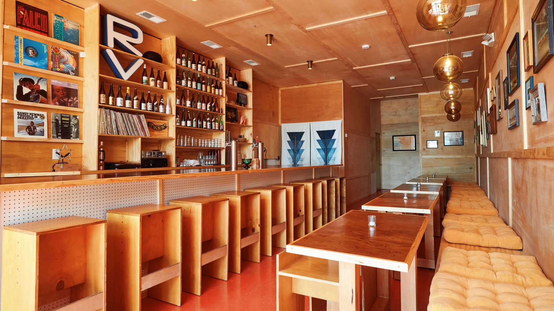 An empty sake bar is seen from an angle. The bar stools, bench seating and back bar are all built of light wood, and the back bar is filled with sake bottles and records.