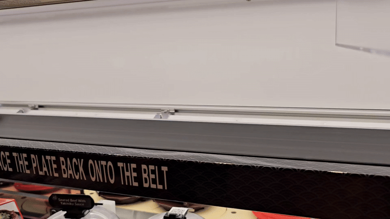 Close-up of a conveyor belt sushi system with an instruction saying "PLACE THE PLATE BACK ONTO THE BELT." Plates with sushi dishes appear below the belt in a restaurant.
