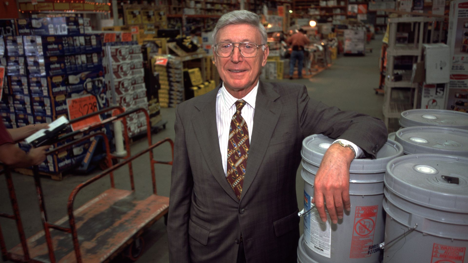 An individual wearing a suit leans against a stack of gray buckets in a large home improvement warehouse store
