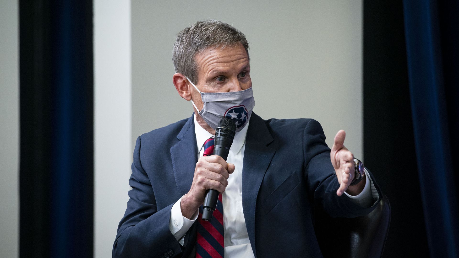 Photo of a masked Bill Lee speaking into a mic and gesturing with his left hand