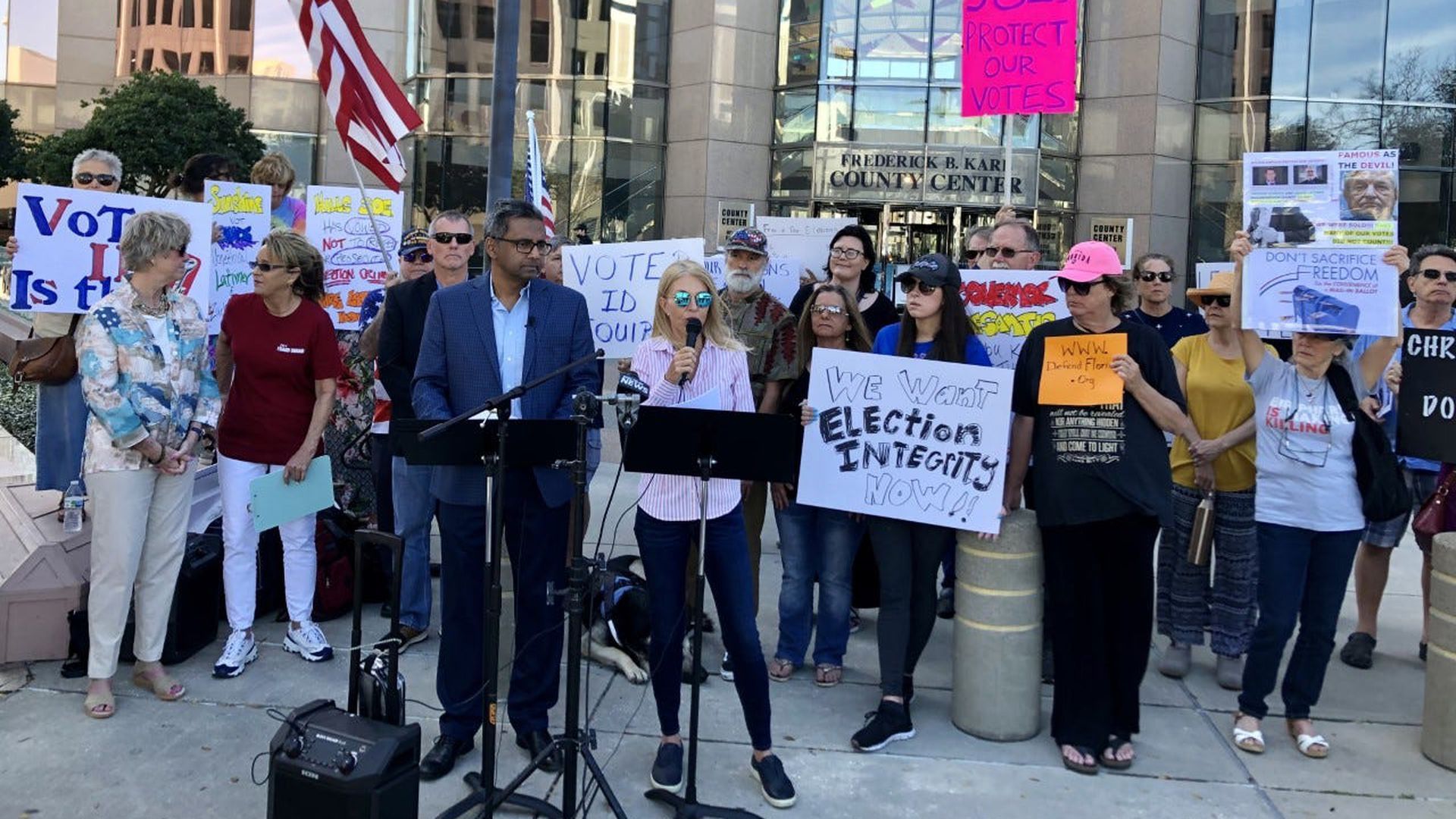 A group of people stand with signs while a woman speaks at a podium