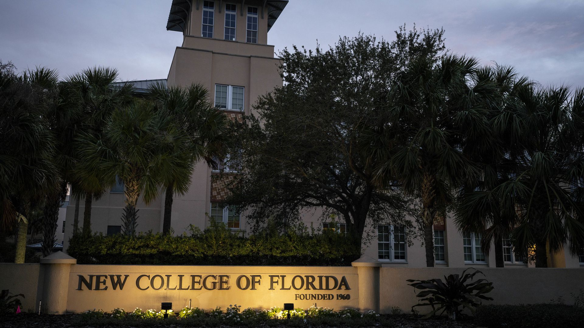A brown building pictured at dusk with lights shining on a sign that says "NEW COLLEGE OF FLORIDA."