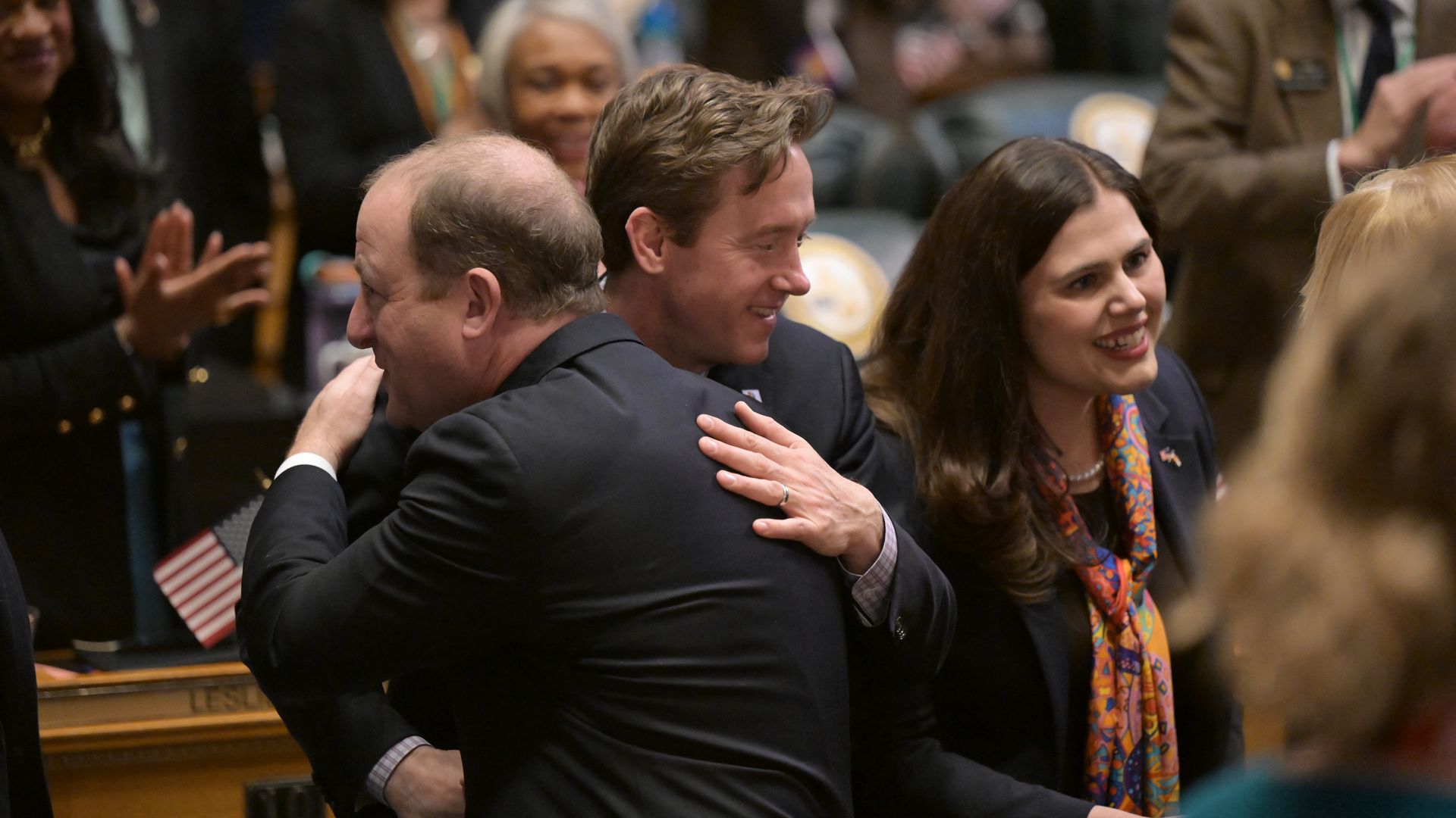 DENVER, CO - JANUARY 11 : Colorado Gov. Jared Polis, front, hugs Denver mayor Mike Johnston by Colorado Secretary of State Jena Griswold, right, before delivering the 2024 state of the state address to a joint session of the legislature in the House chamber at the State Capitol in Denver, Colorado o