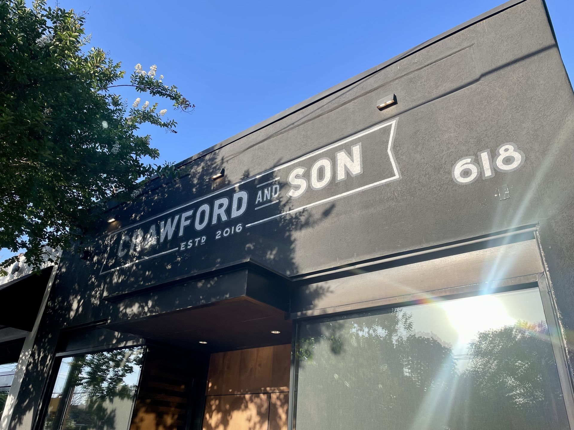 Grey storefront wall with a white-outlined sign reading "CRAWFORD AND SON" and the number 618 to the right; tree branches with white blossoms against a blue sky.