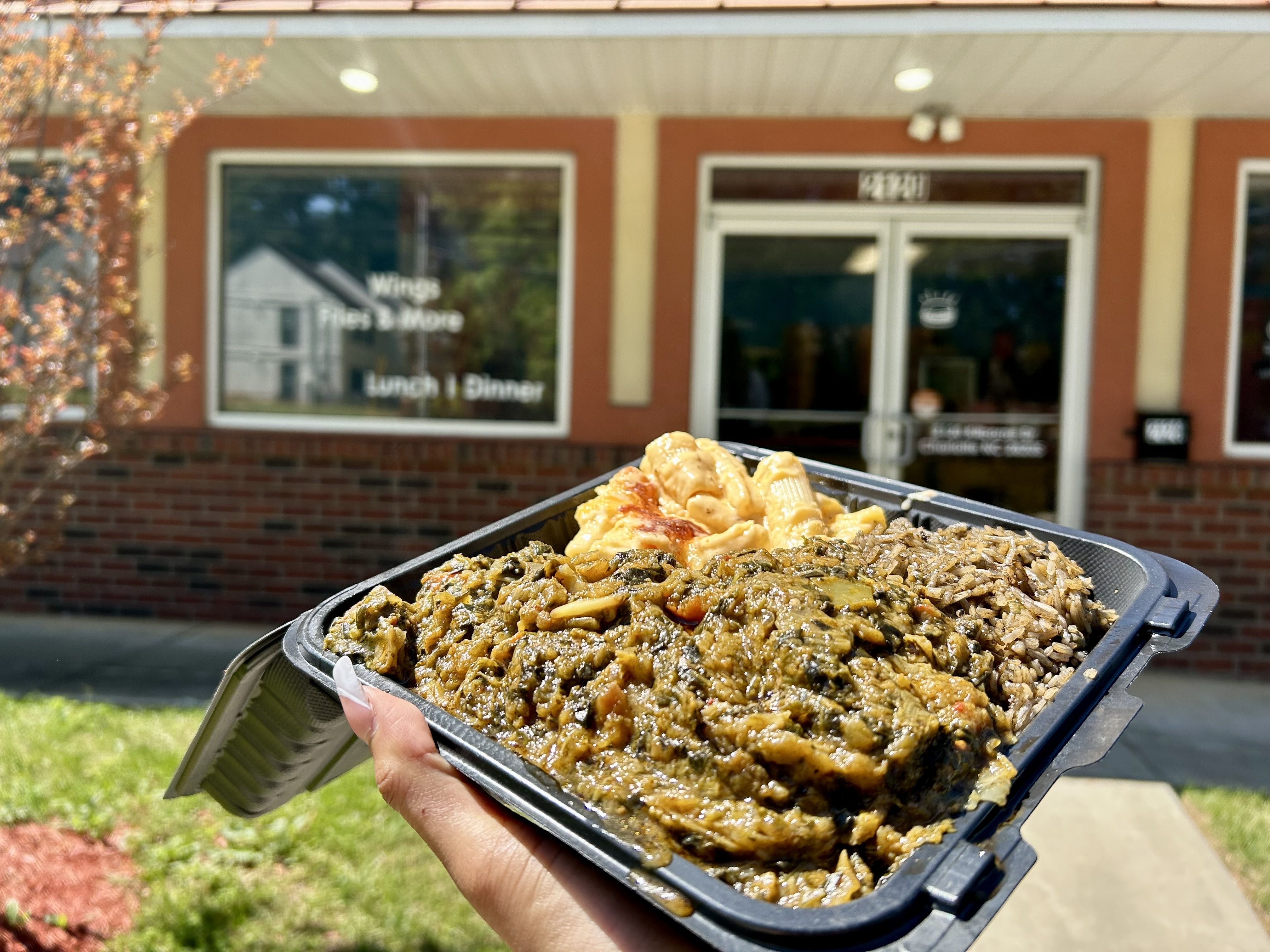 Hand holding a black plastic takeout container with greens, macaroni and cheese, and rice in front of a brick building with a glass door and window showing meal options like wings and fries.