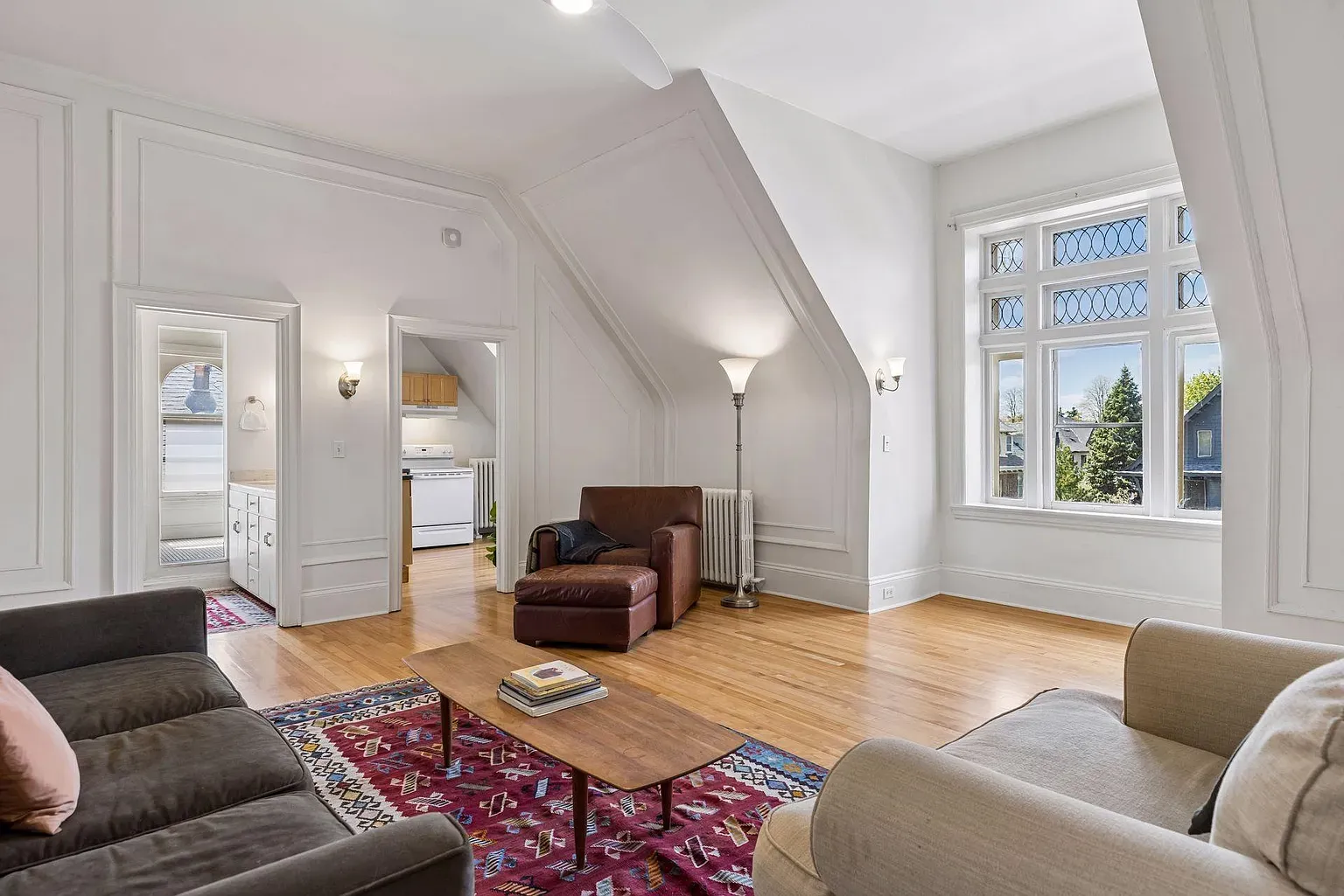 Bright living room with white paneled walls and a sloped ceiling. A brown leather armchair and ottoman, floor lamp, gray sofa, beige chair, and a colorful rug surround a large bay window and a doorway