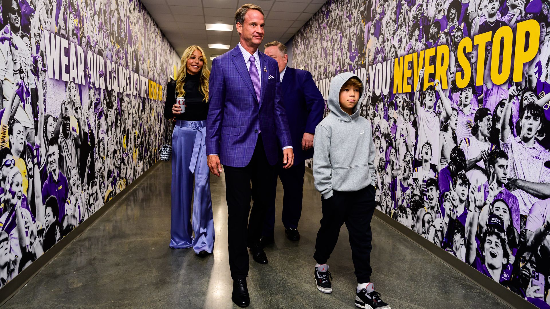 A man walks ahead of a young boy, woman and another man through a hallway. The walls are decorated with gray-toned fan photos with purple and yellow accents.
