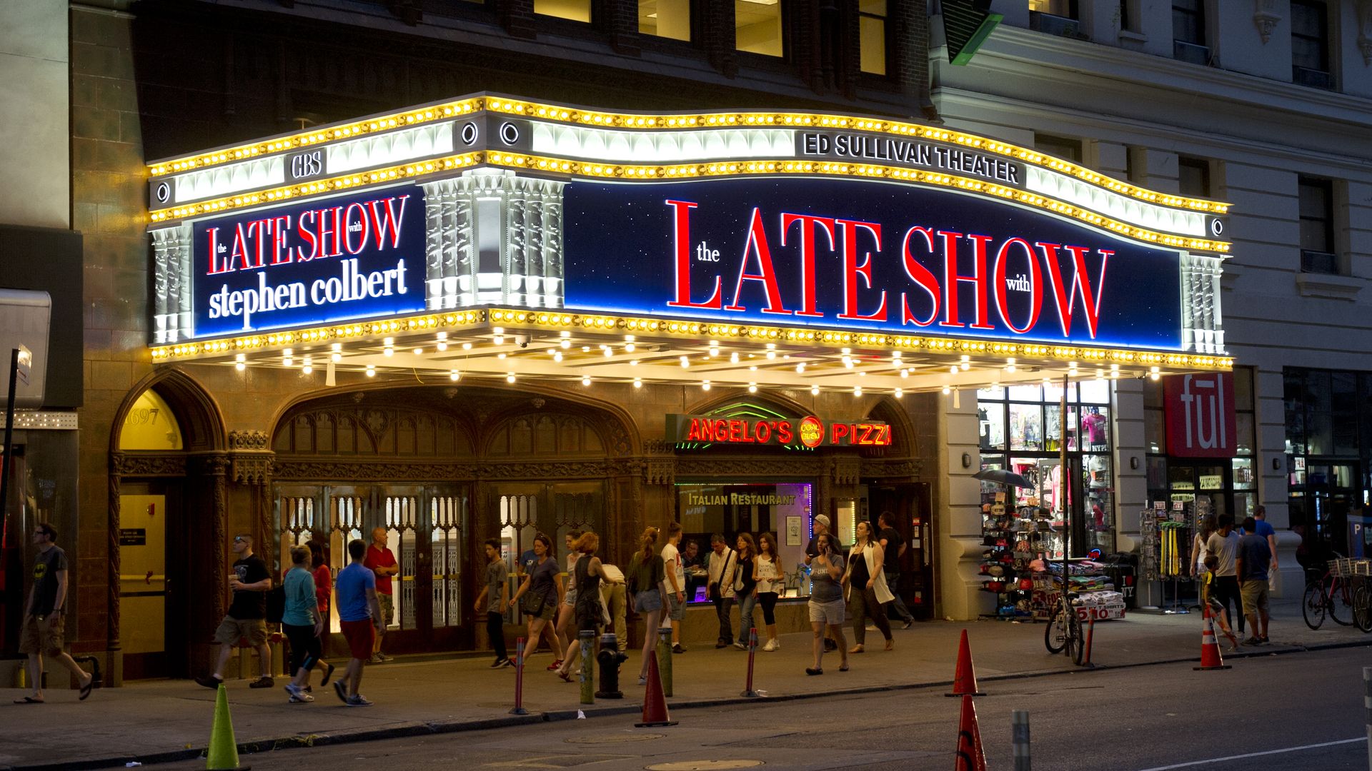 Nighttime street scene in front of the Ed Sullivan Theater marquee advertising "The Late Show with Stephen Colbert"; bright yellow bulbs frame bold red lettering as people walk below.