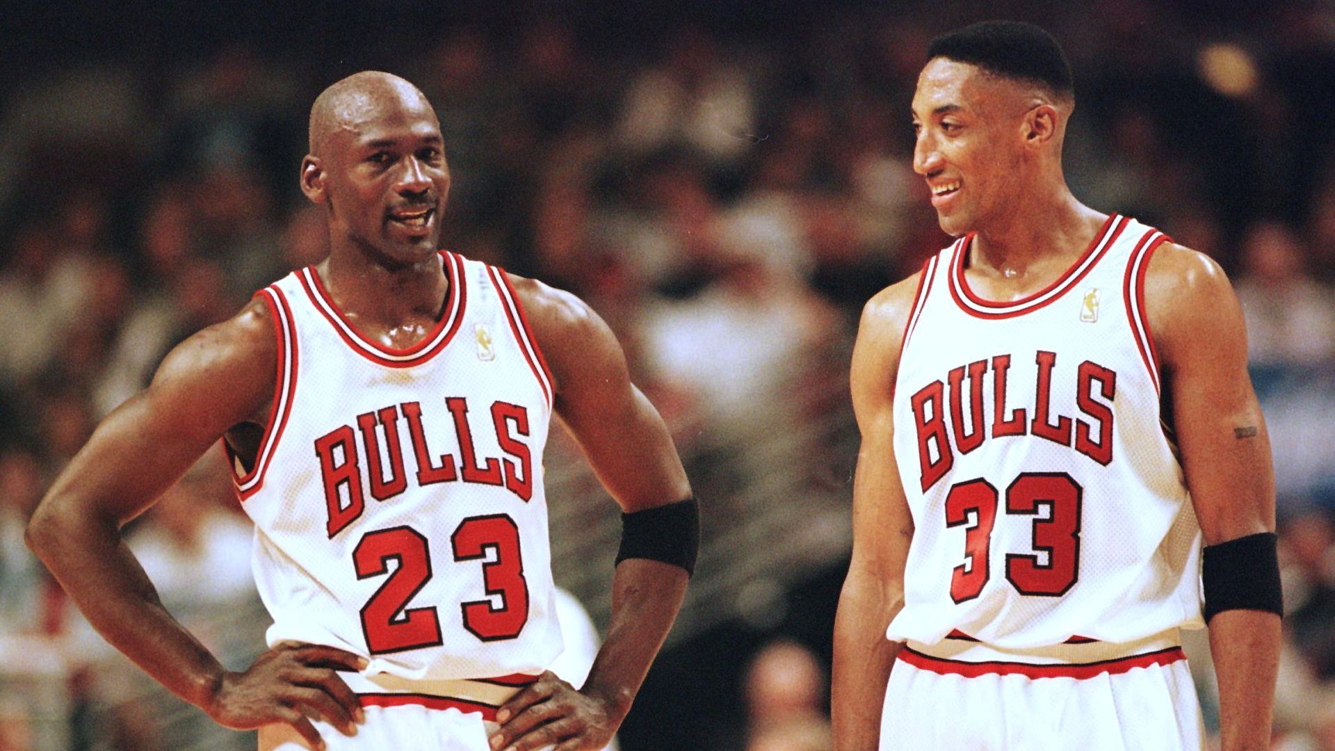 Two Chicago Bulls players in white jerseys with red trim, numbers 23 and 33, stand on a basketball court with hands on hips, smiling, as a blurred crowd watches from the stands.