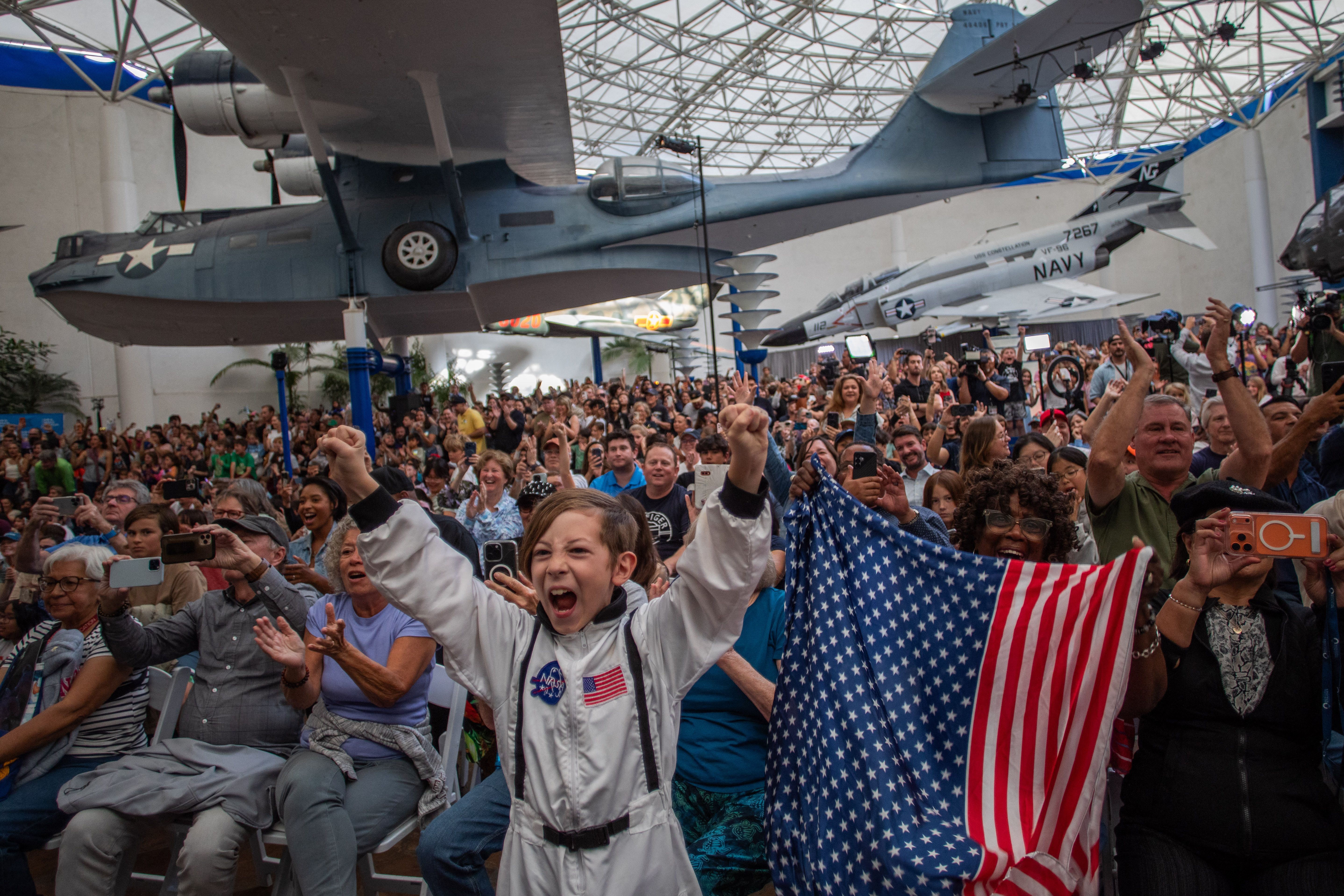 A young boy in an astronaut costume stands next to a woman waving a flag as they watch a live broadcast of the Artemis II crew’s return during a watch party at the San Diego Air and Space Museum in San Diego, California, on April 10, 2026