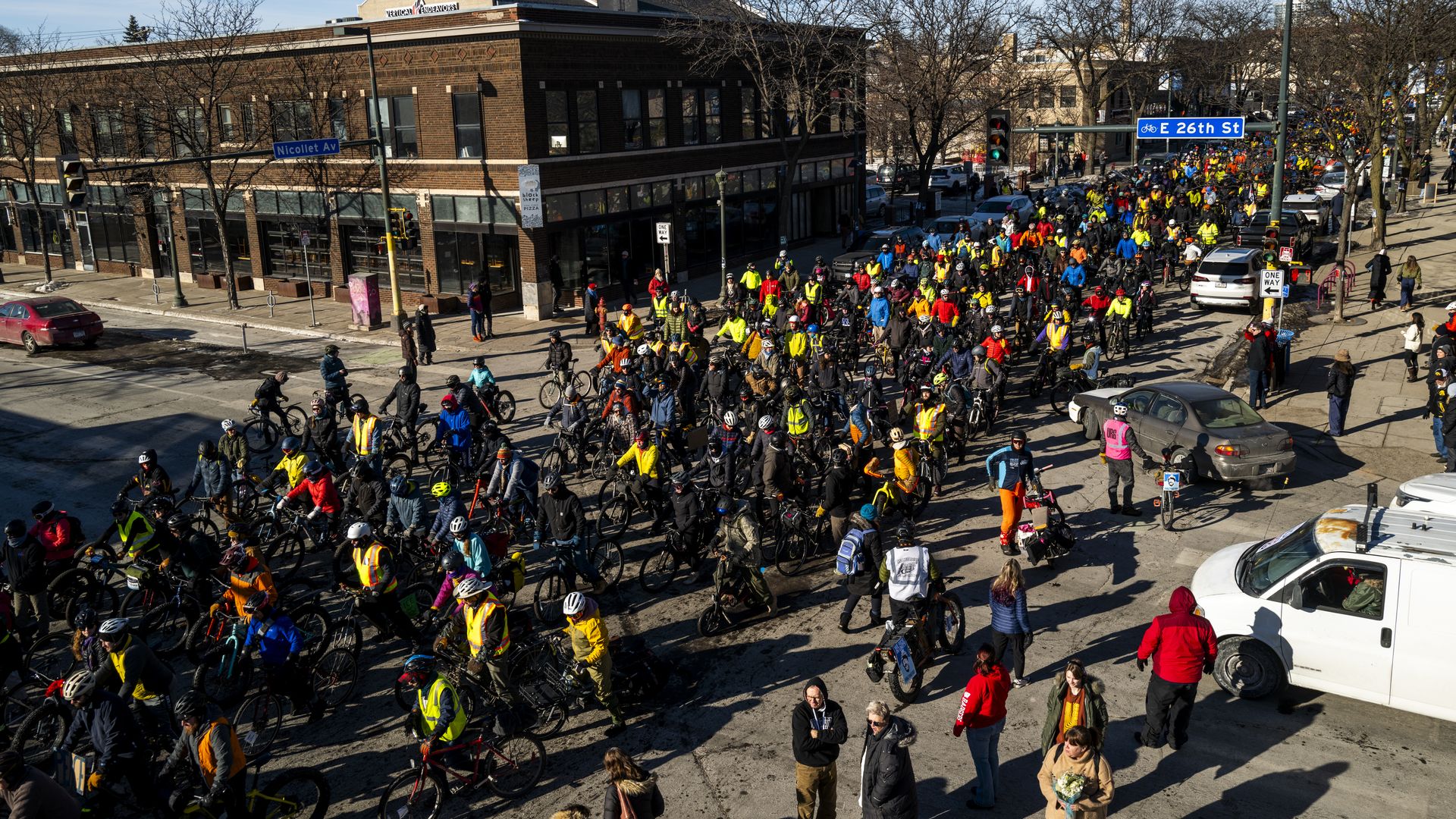 Hundreds of people on bicycles ride through an intersection with a 26th Street sign above them and brick building in the background. 