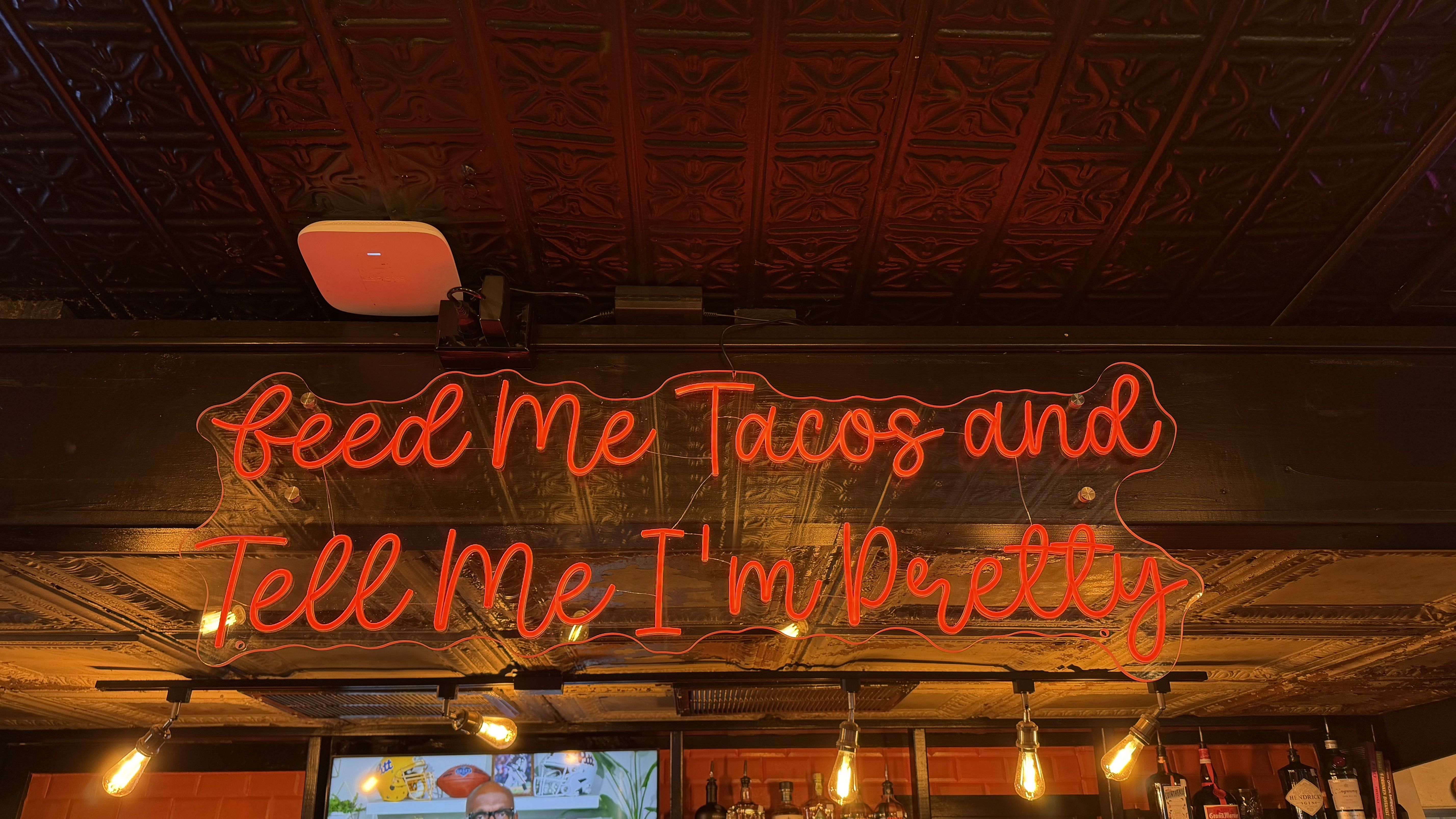 Bar interior with warm lighting and a red neon sign reading "Feed Me Tacos and Tell Me I'm Pretty" above bottles; ornate dark tin ceiling, exposed bulbs, and a white wall-mounted device on the left.