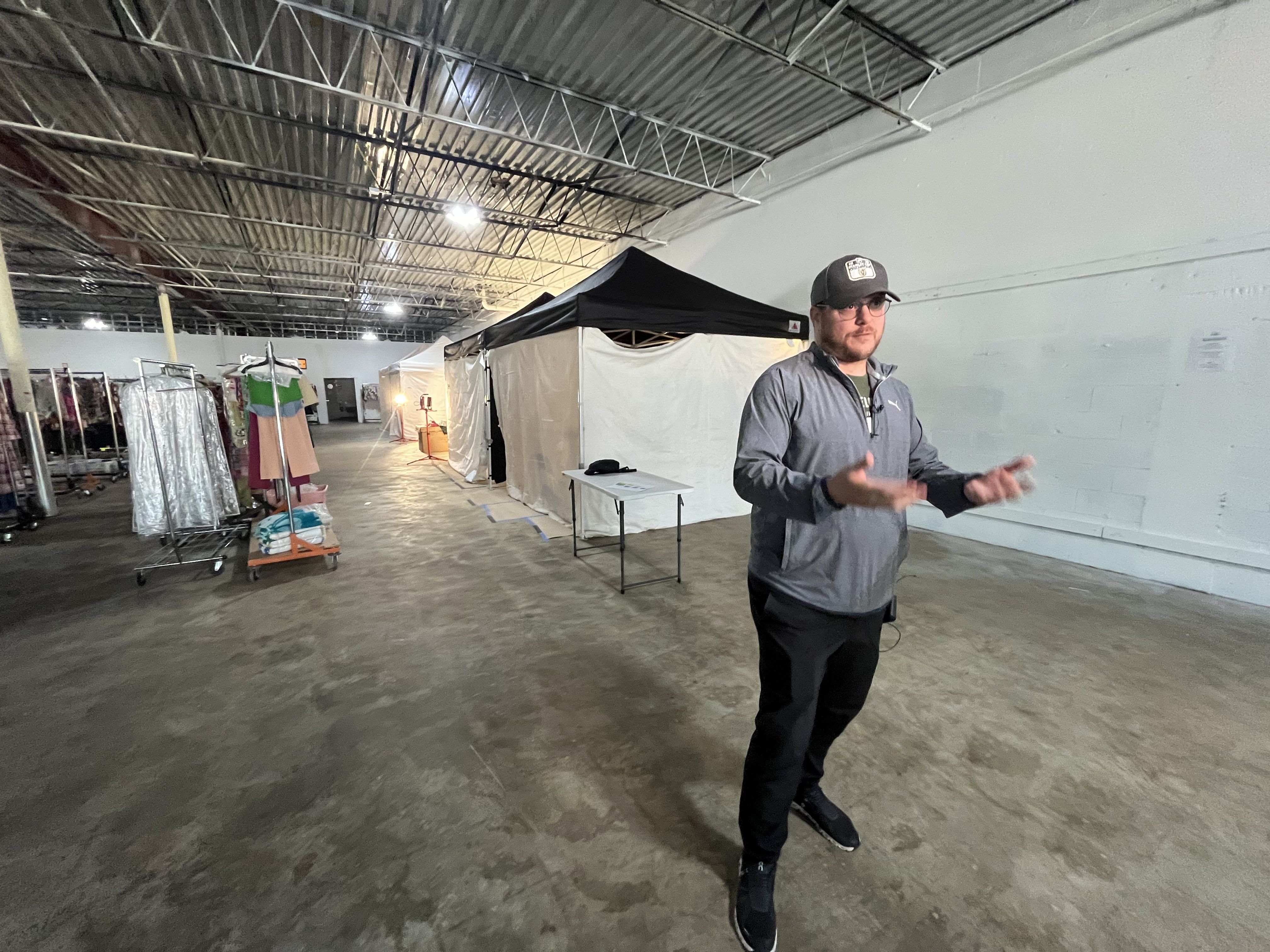 A man in a gray zip-up jacket and baseball cap stands in a large open warehouse with a concrete floor, white tents, and clothing racks on the left. He gestures as if presenting.