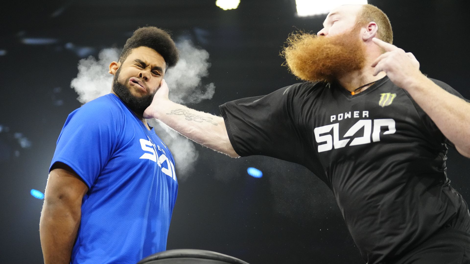 A man grimaces as he holds his hands behind his back and is slapped in the face by another man.