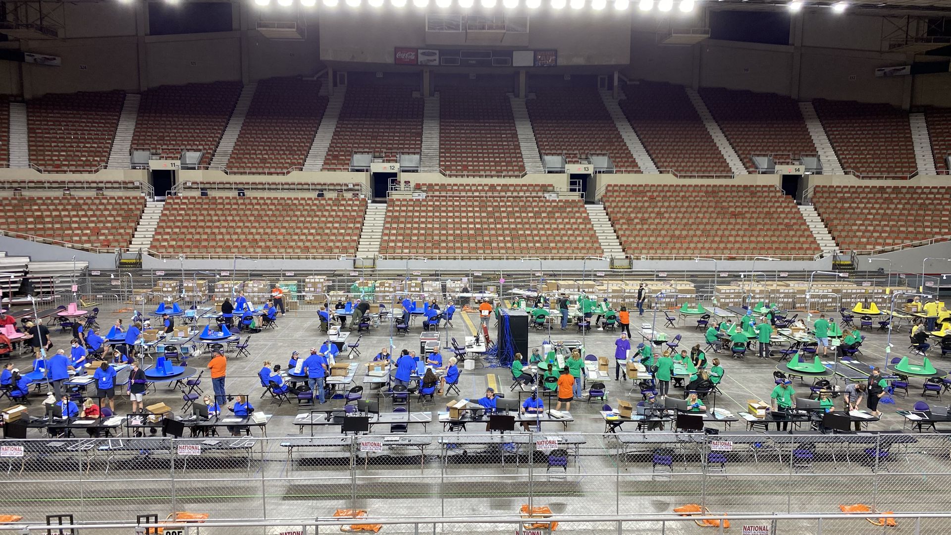 Red, blue, green and yellow tables on the floor of an arena, staffed by people wearing shirts of the same colors.