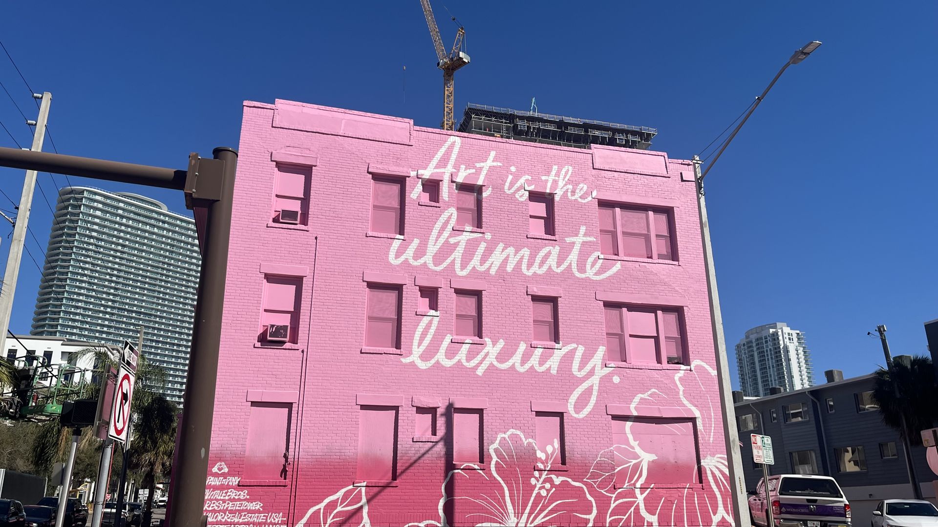 Bright pink building with white cursive text reading "Art is the ultimate luxury," surrounded by white floral designs under a clear blue sky in an urban area.