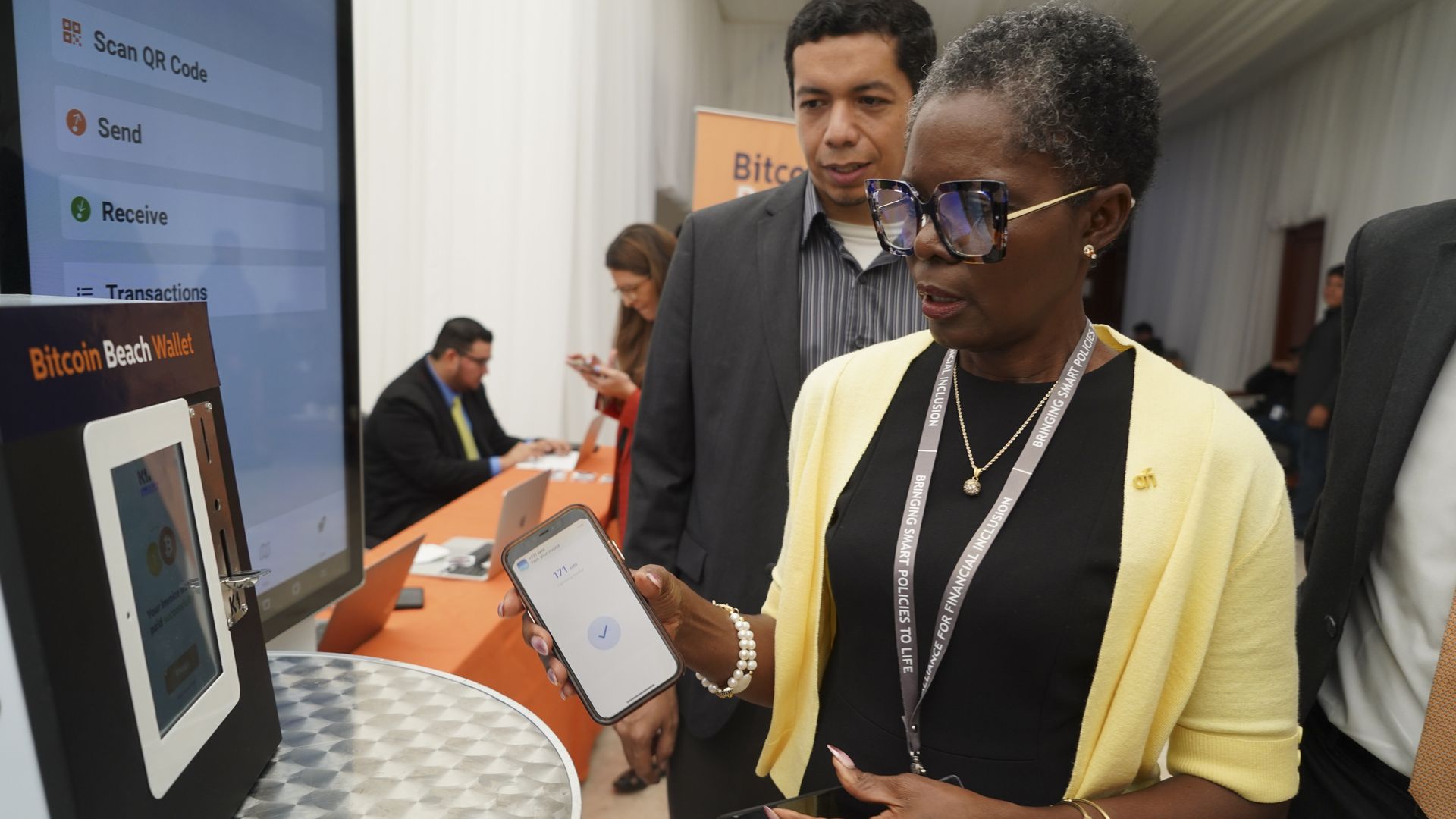 a woman holds her phone to a computer screen while learning about bitcoin