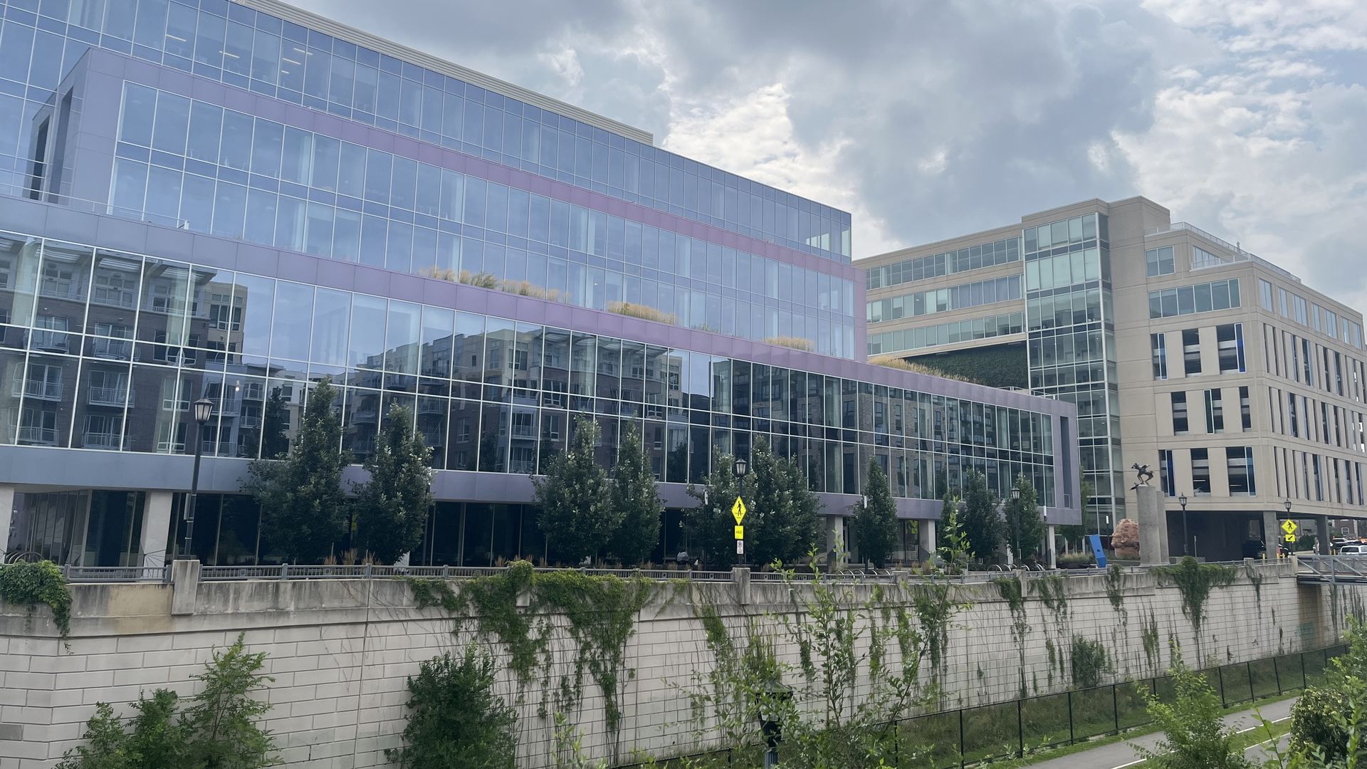 Modern glass and concrete buildings with green trees in front, a cloudy sky above, and a pedestrian path below a concrete wall with climbing ivy.