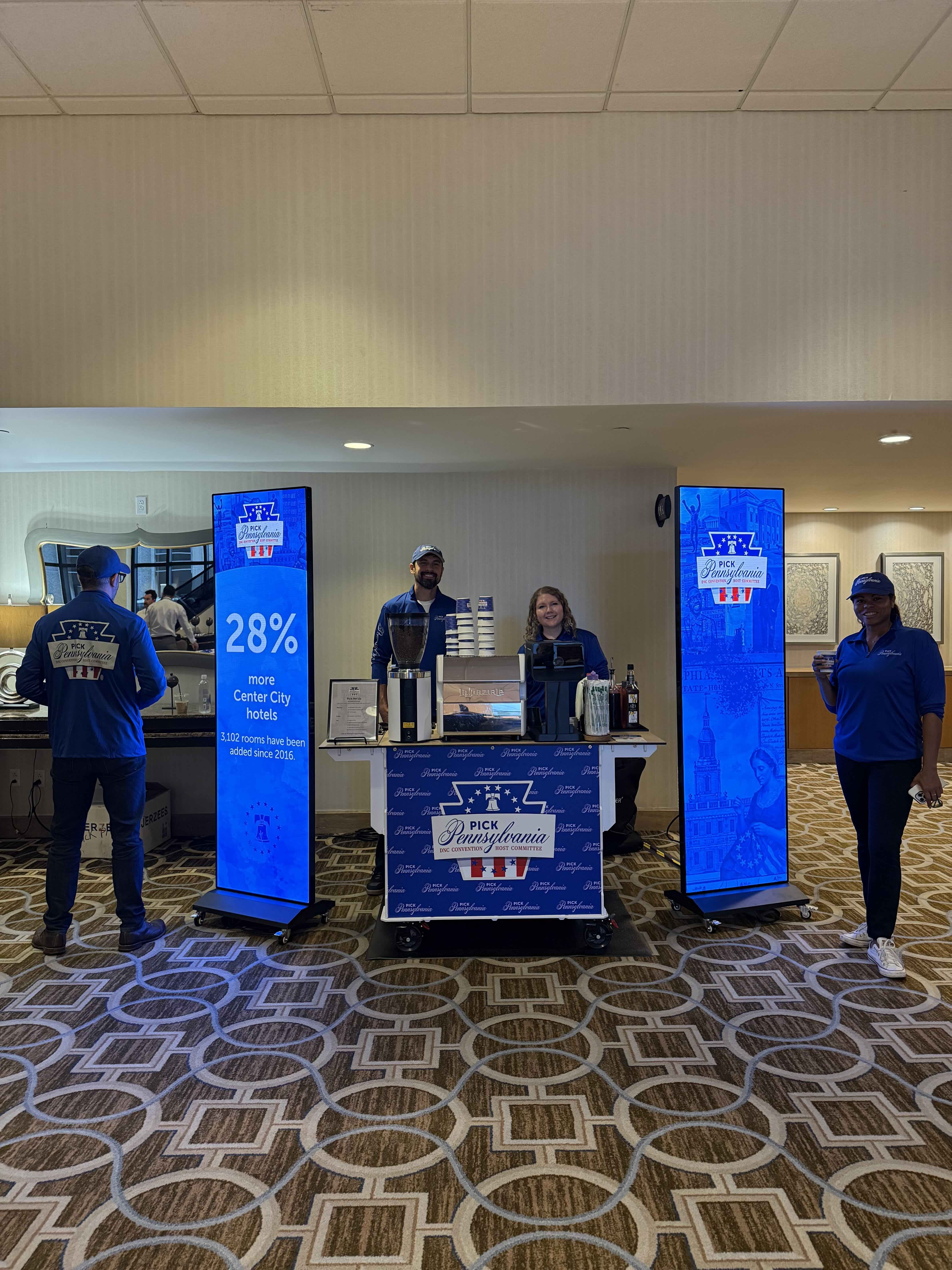 Indoor convention booth with two tall blue digital displays beside a central counter labeled "Pick Pennsylvania". Three staffers in blue shirts stand behind the counter; patterned carpet and ceiling tiles visible.