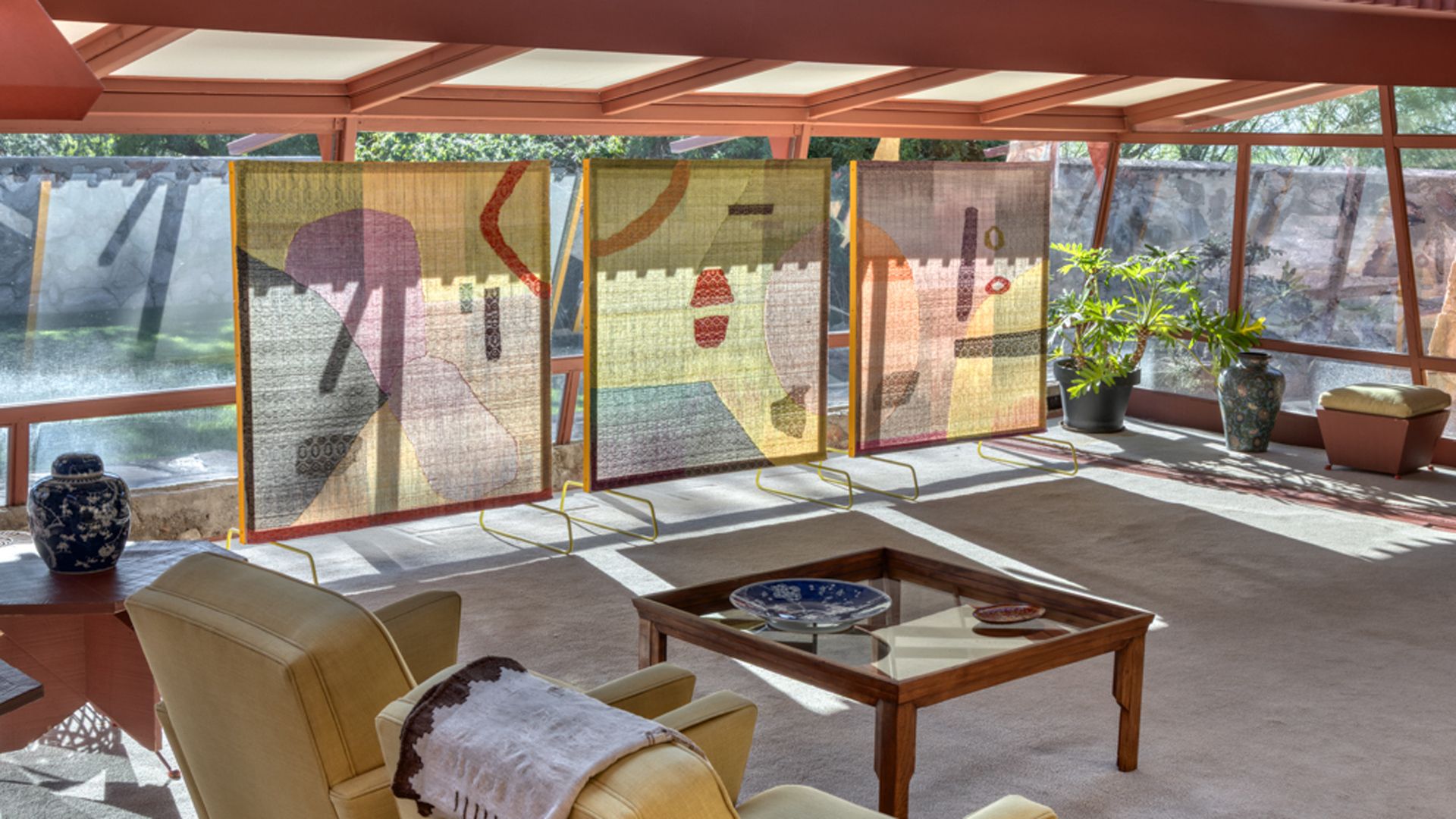 Sunlit room with three colorful abstract panels on stands, beige armchairs, a wooden glass-top coffee table, patterned vases, green plants, and large windows showing outdoor stone wall.
