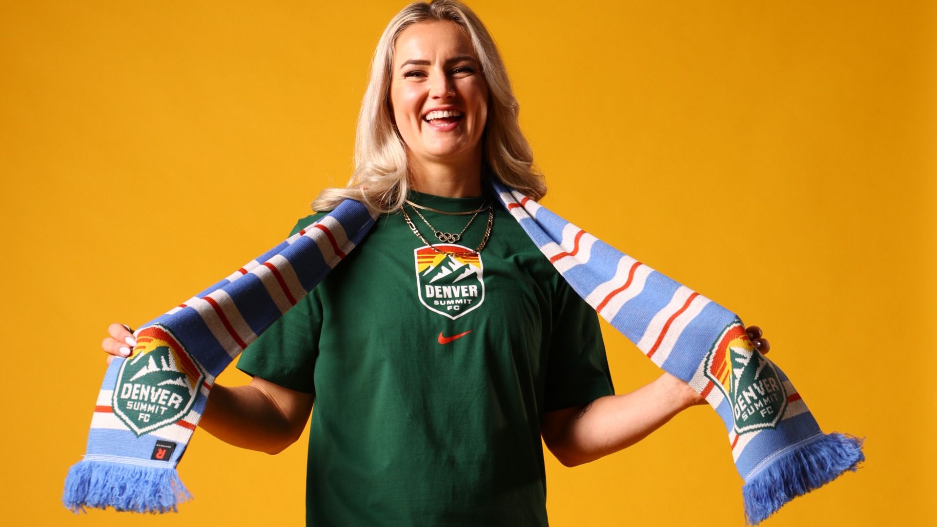 Smiling blonde woman wearing a green "Denver Summit FC" shirt and holding a blue, white, and red striped scarf with the same logo, against a solid orange background.