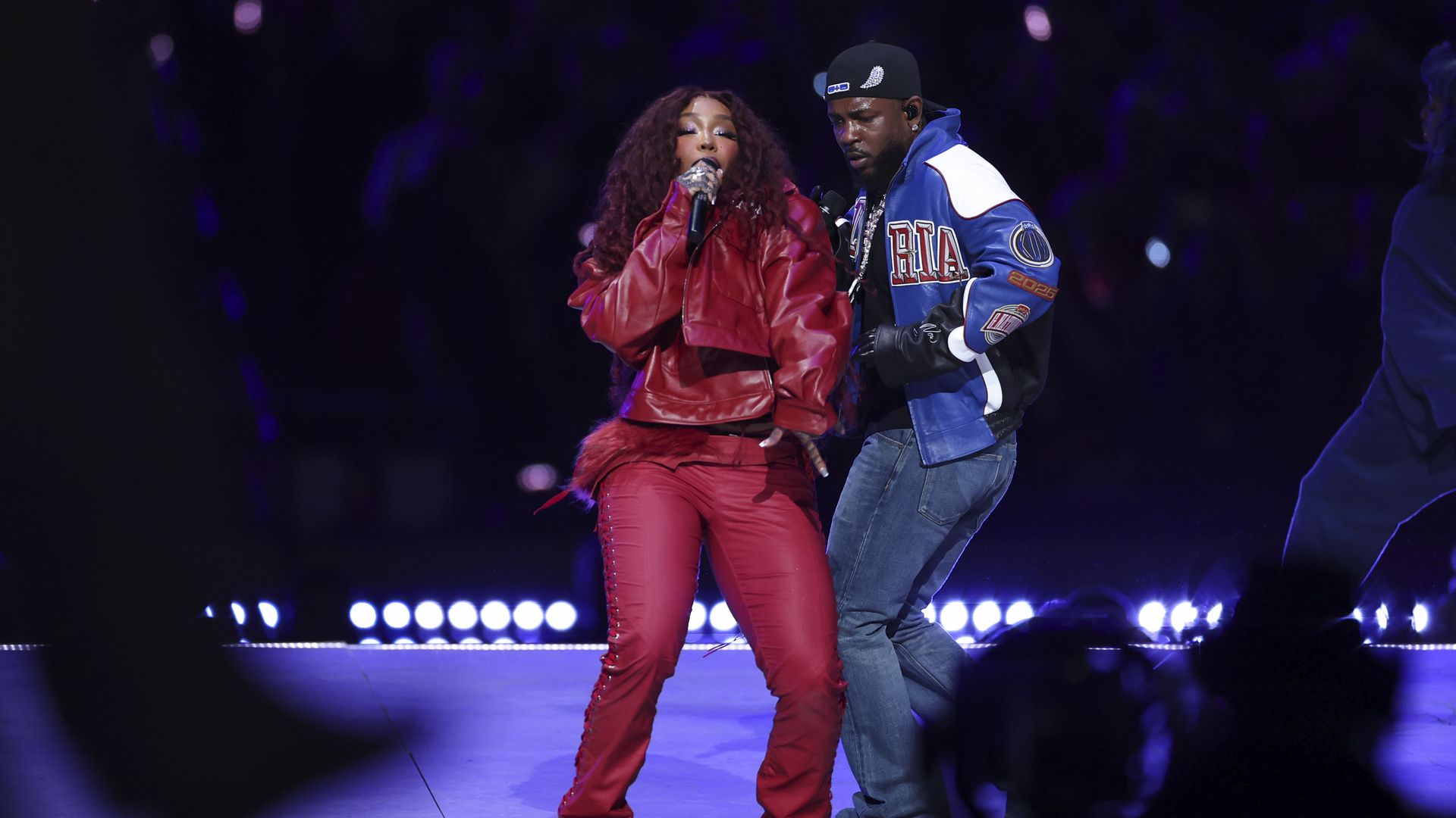 Two individuals — one wearing a dark red outfit, the other dressed in jeans and a leather sports jacket — perform on stage during a Super Bowl halftime show