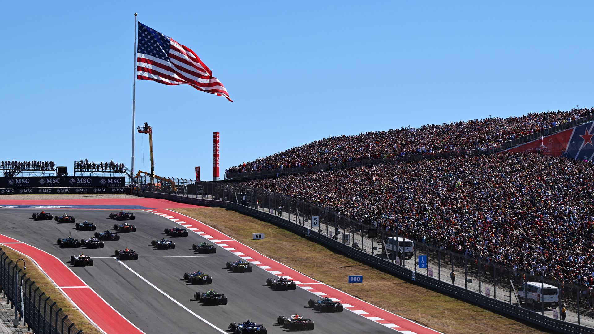 Wide shot of a Formula 1 race on a curved track, with a large waving American flag on a tall pole left, a packed grandstand to the right, and many race cars speeding under a clear blue sky.
