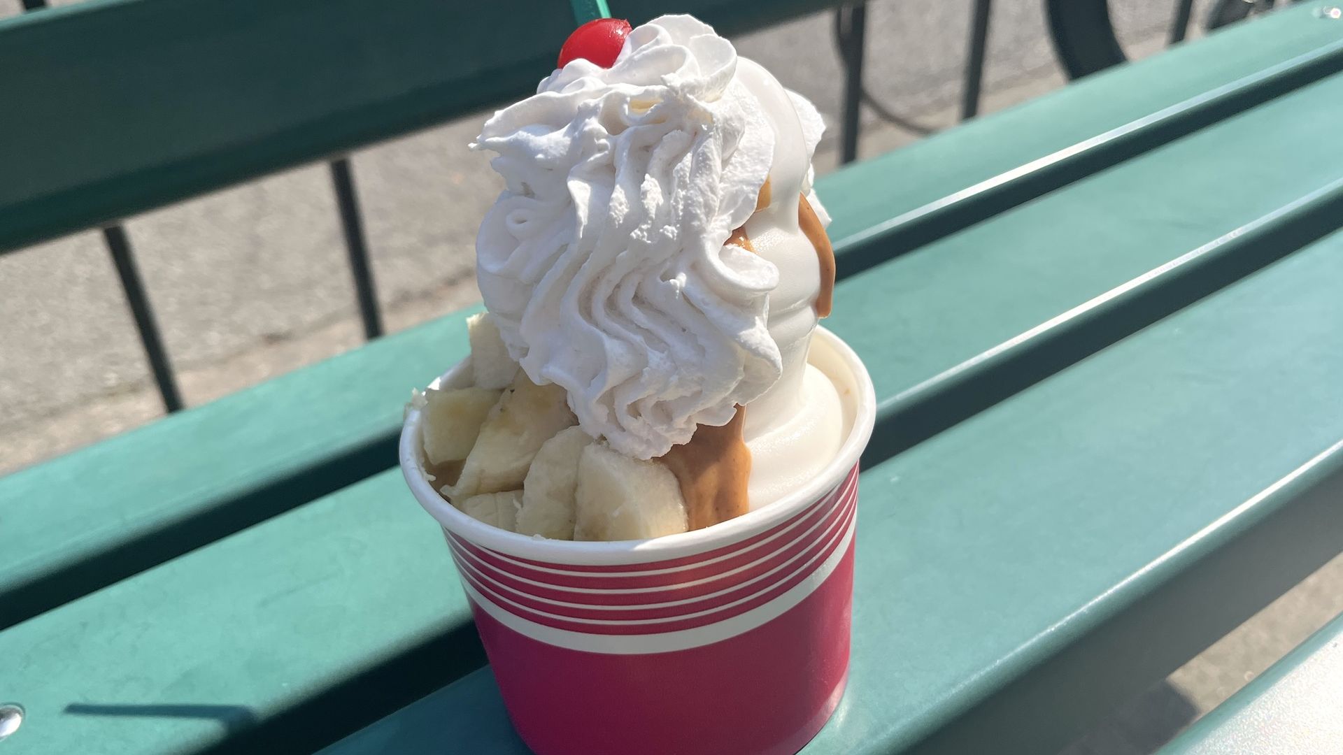 Pink cup with bananas, ice cream, peanut butter sauce, whipped cream and a cherry sitting on a green bench.