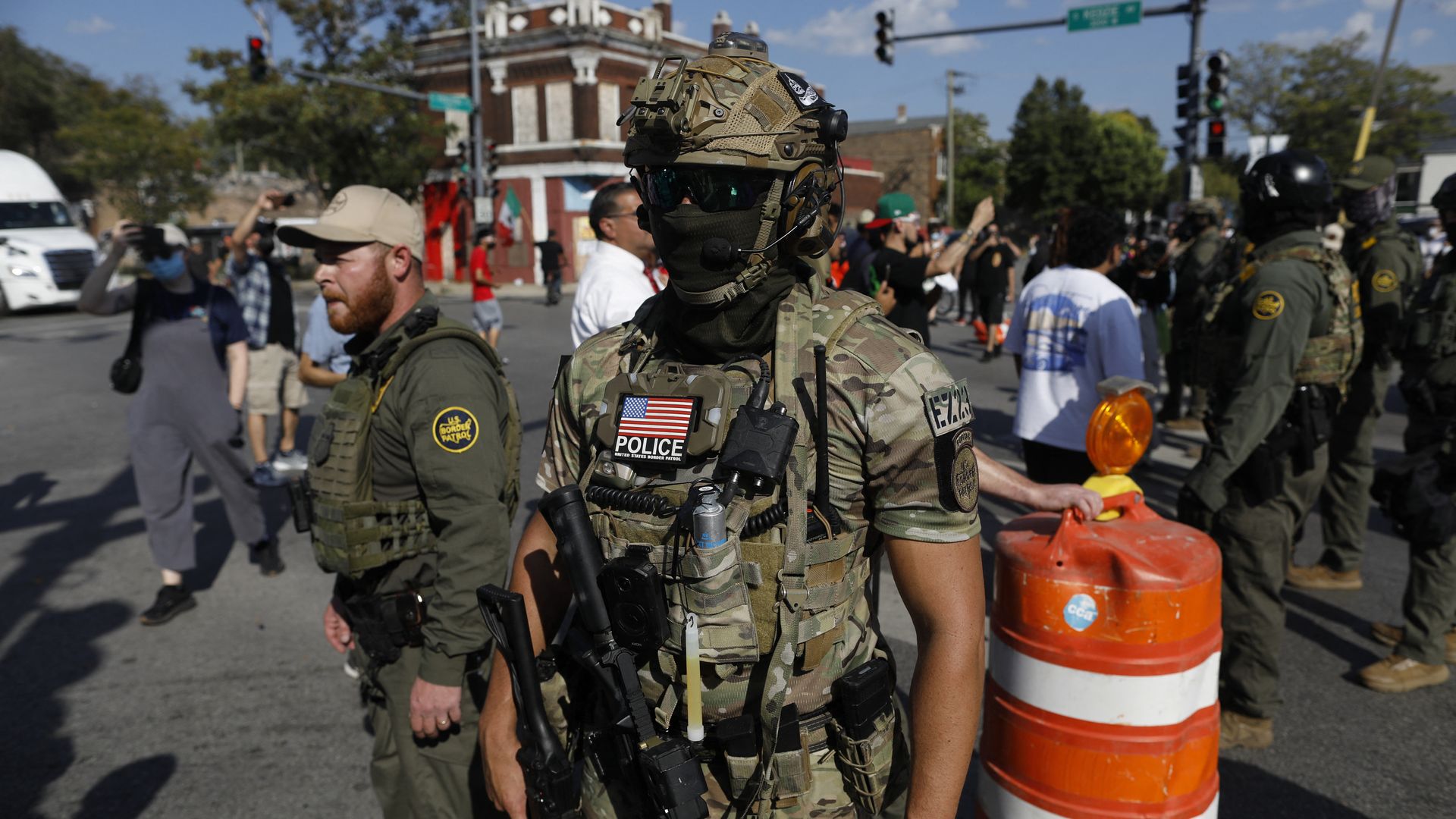 A masked U.S. Customs and Border Protection agent in front of an orange barrier with other officers and residents on a Chicago city street.