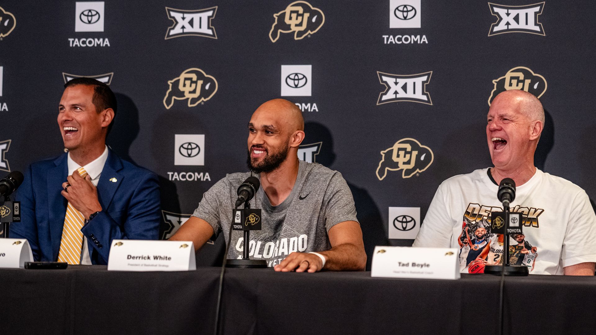 Three men sit at a press conference table in front of a dark backdrop with logos. Microphones visible; left man in blue suit laughs, middle man gray shirt smiles, right man white T-shirt laughs.