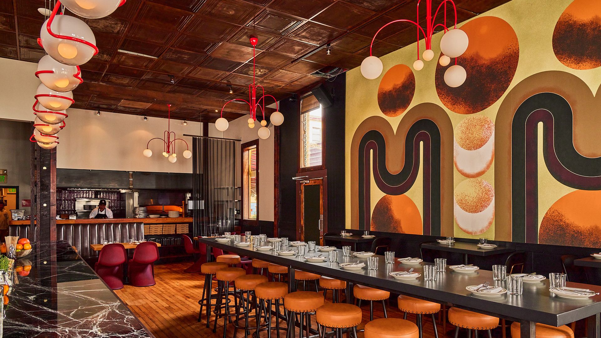 Interior of a restaurant with warm wood floors, a long black table with orange stools, retro red and white light fixtures, and a bold wall mural featuring brown, orange, and black geometric shapes.