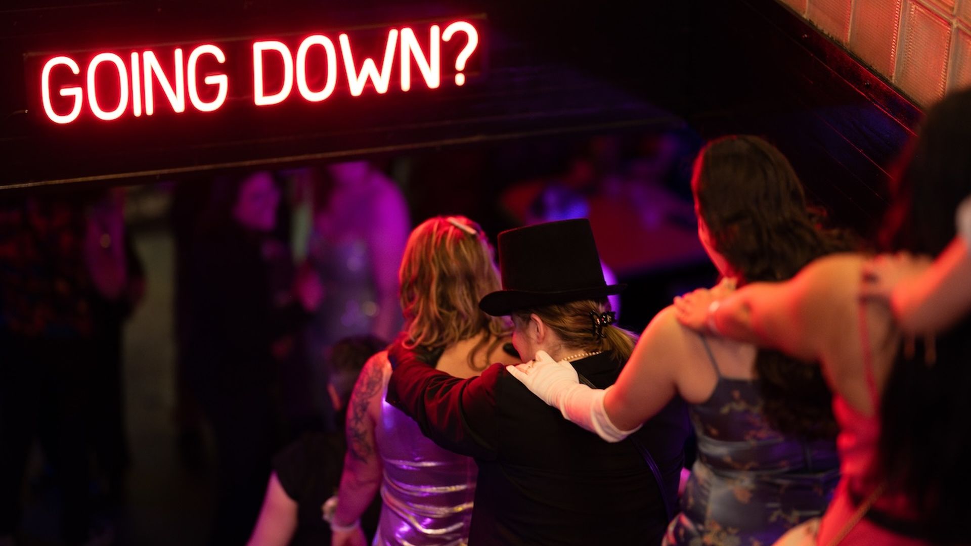 A group of people dressed in formal attire and party clothes descending stairs, illuminated by a neon sign reading "GOING DOWN?" in a festive and dimly lit setting, hinting at an event or gathering.
