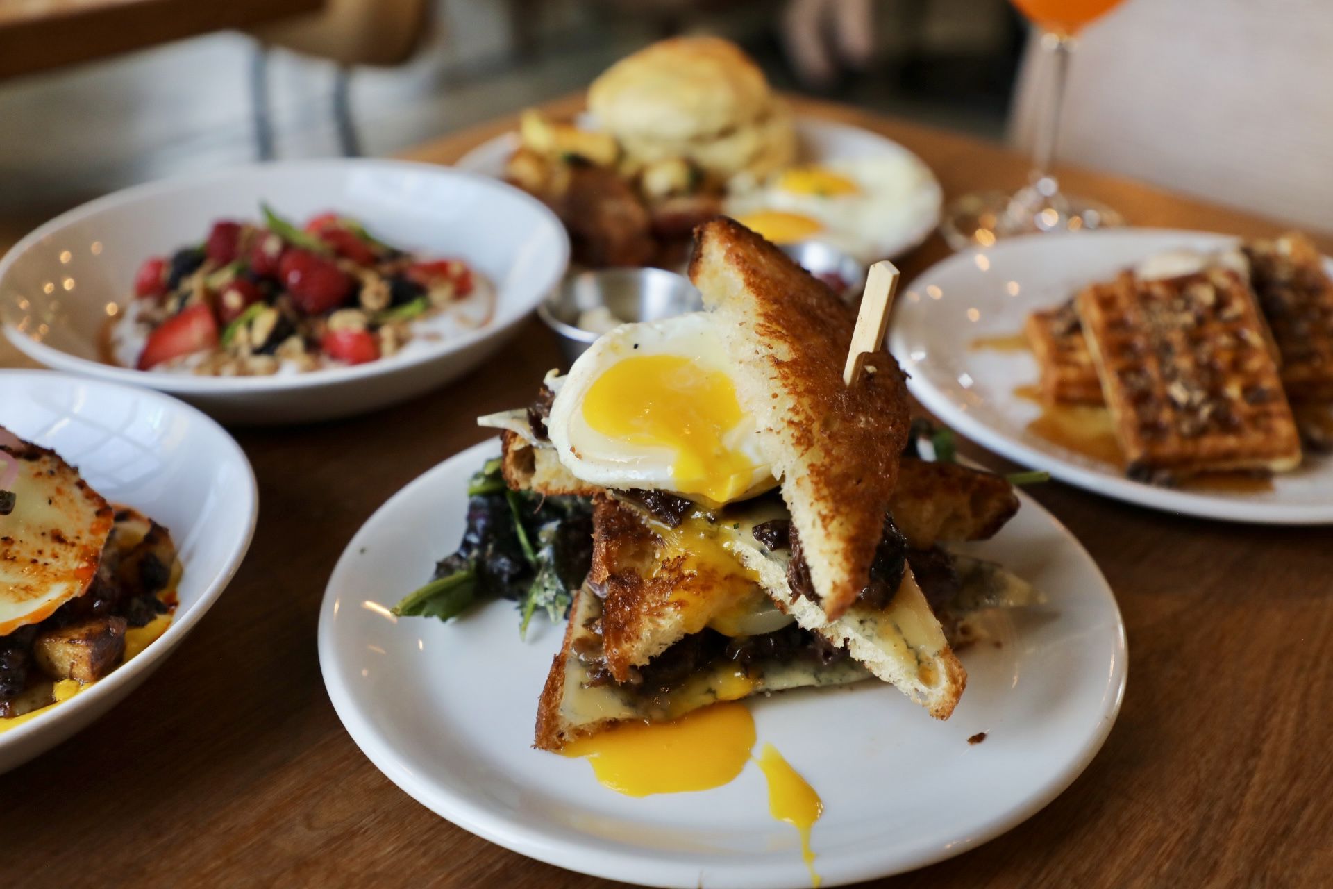 Brunch plate with a stacked grilled sandwich topped by a runny egg, melted cheese, and greens. In the background: a berry yogurt bowl, waffles, and an orange drink.