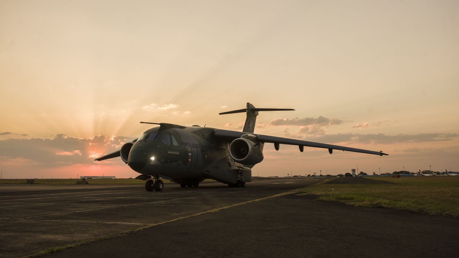 Olive-green military plane on a runway at sunset, with orange sky and sun rays.