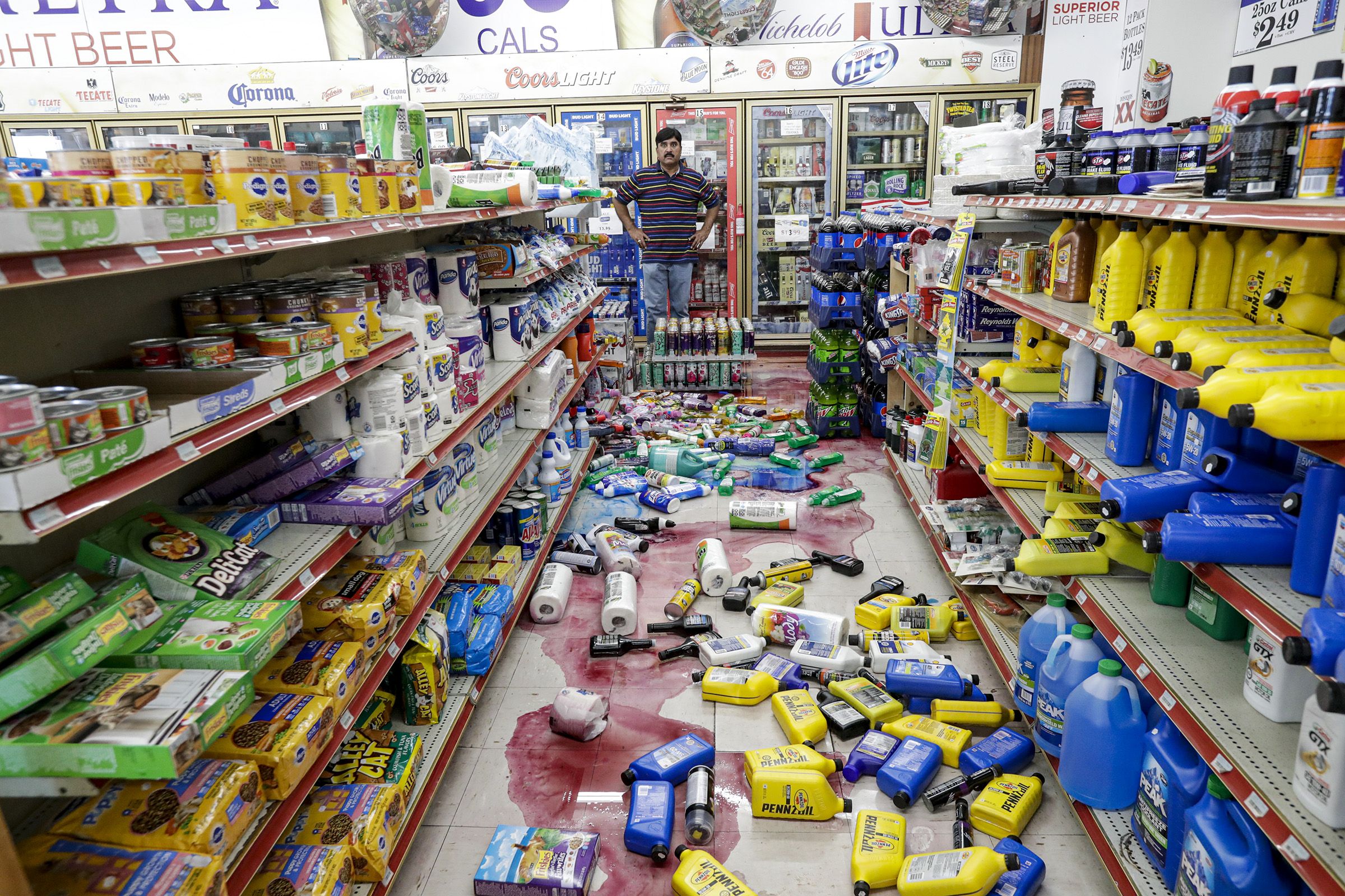 This image shows a man standing in a grocery store aisle covered in spilling bottles.