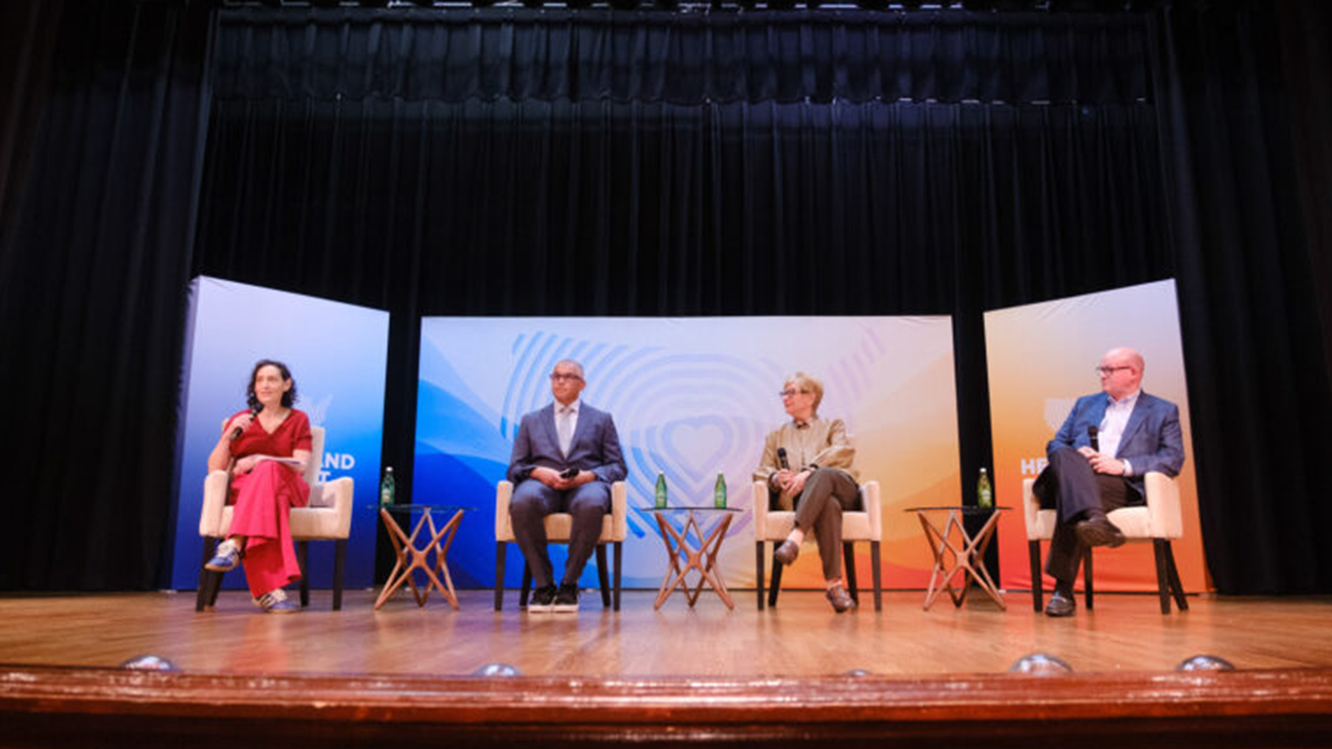 Panel discussion on a stage with four speakers in beige chairs, blue/orange backdrop panels, small side tables, water bottles, and dark curtains.