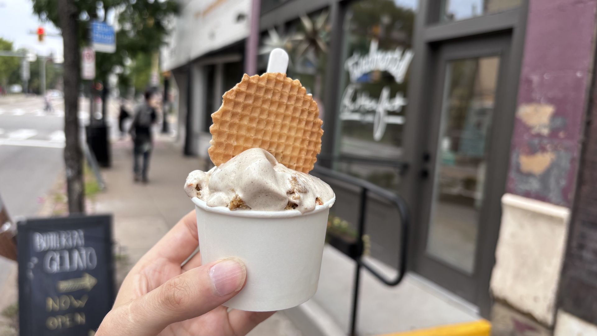 Close-up of a hand holding a white cup of cookies and cream ice cream topped with a round golden waffle wafer, set against a blurred city street with storefronts.