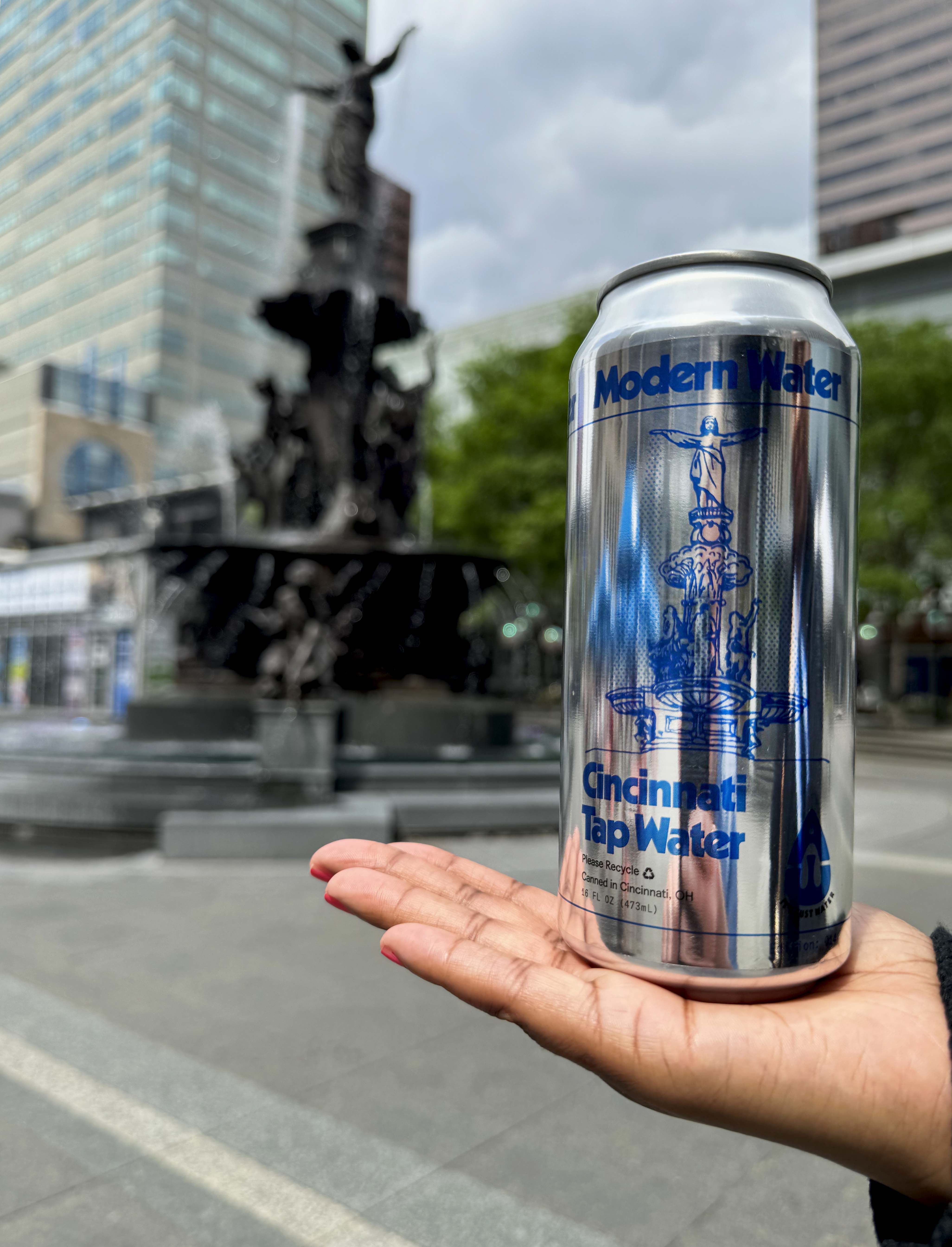 Close-up of a shiny can labeled "Modern Water" and "Cincinnati Tap Water" held by a hand with red nails; a dark statue fountain and glass buildings blur in the background.