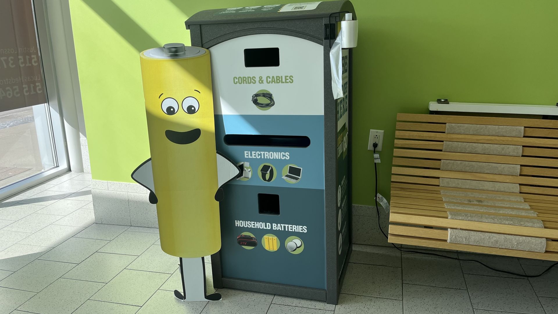 Cartoon yellow battery stands beside a teal-gray electronics recycling bin labeled CORDS & CABLES; lime green wall, tiled floor, and a wooden bench nearby.