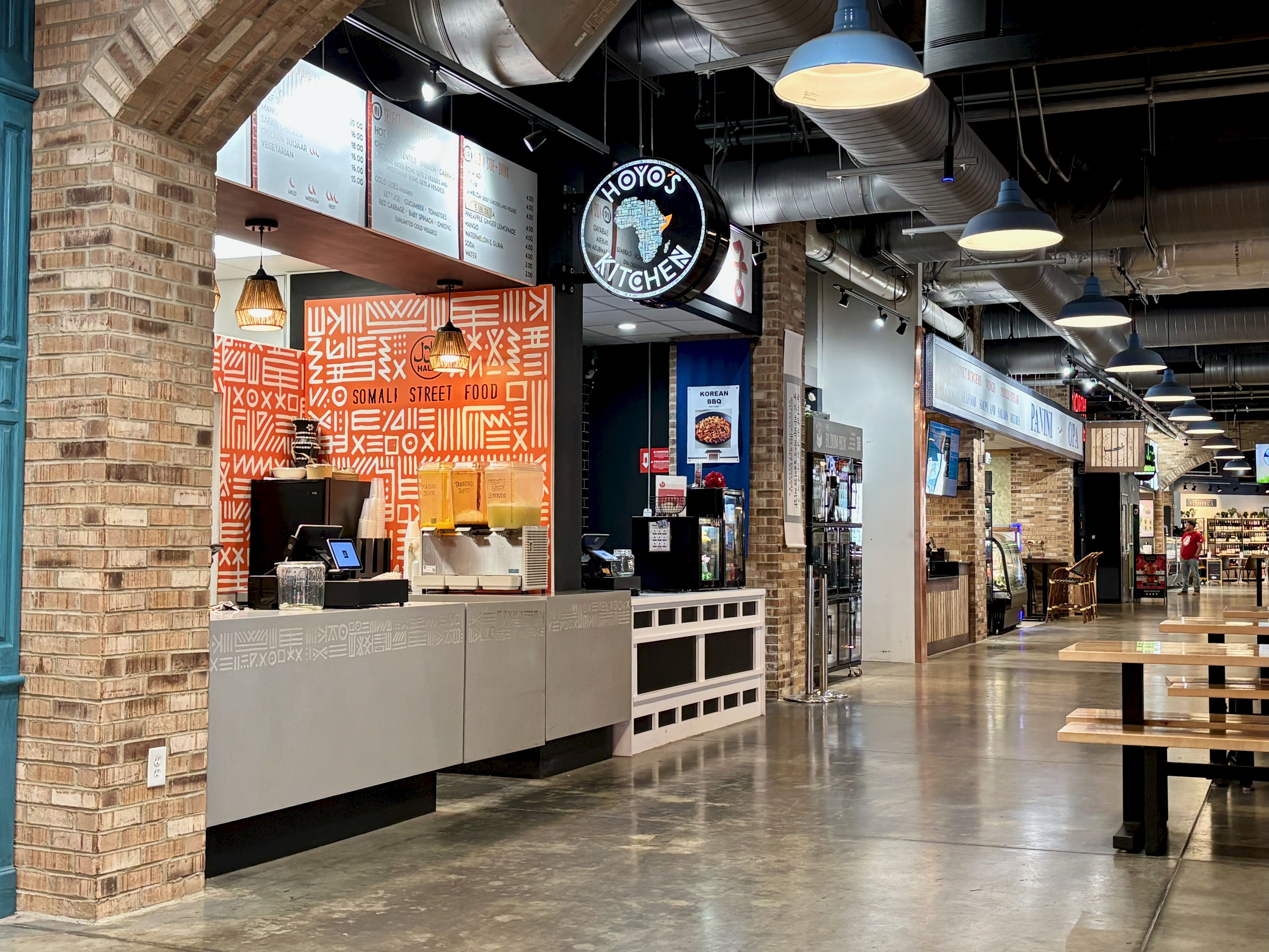 Industrial food hall with brick columns and exposed ducts. A counter fronts an orange geometric wall reading "Somali Street Food". Menu boards, drink station, and blue pendant lights line the hallway.