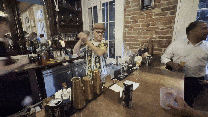 A man in a fedora shakes a cocktail at a busy bar.