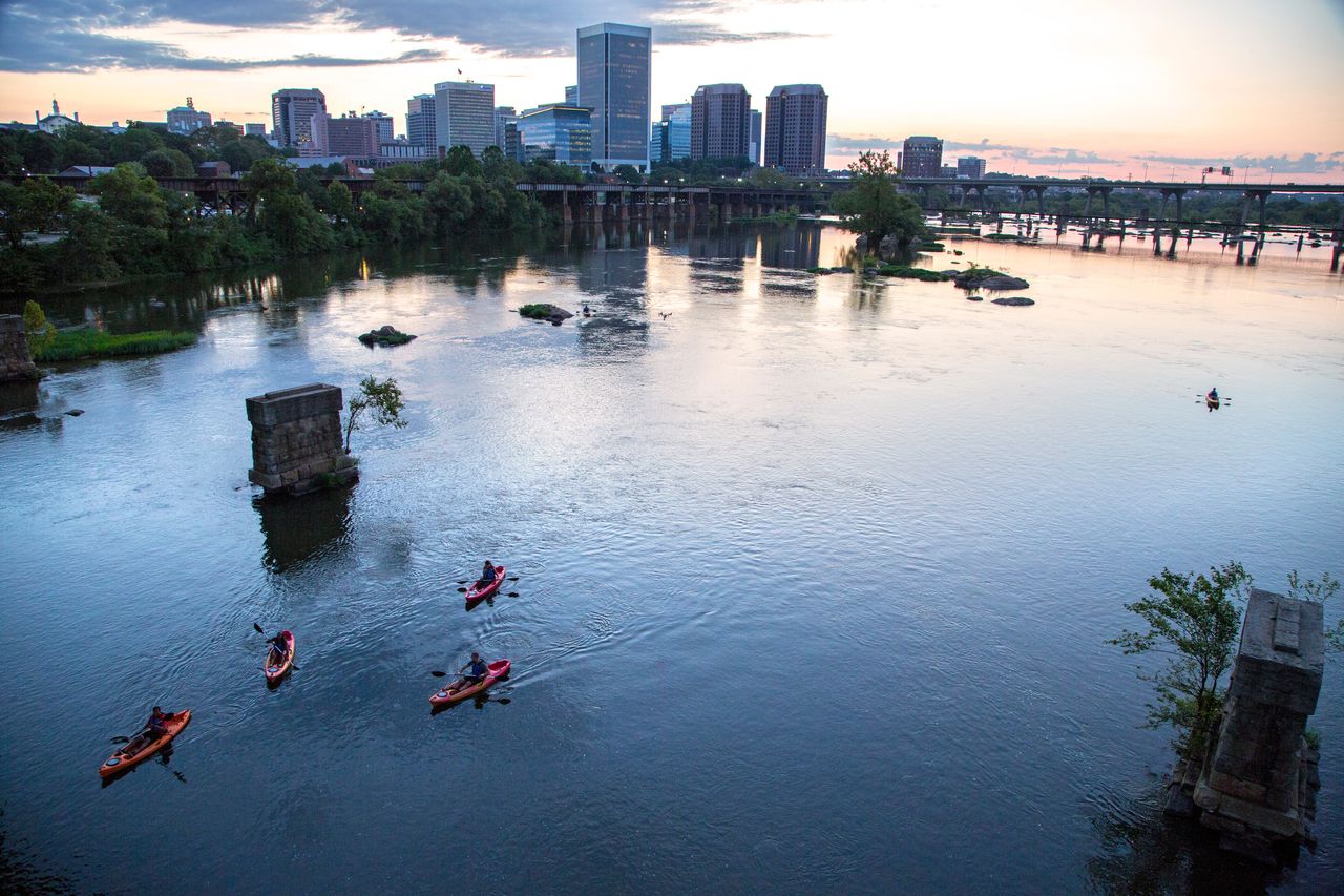 Five people kayaking in red kayaks on a calm river with a city skyline and bridges at sunset under a partly cloudy sky.