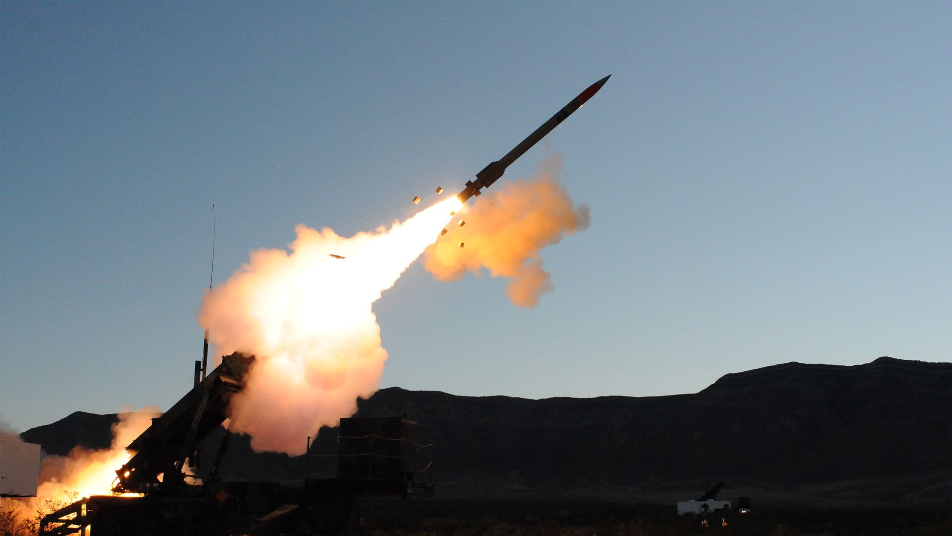 Rocket launches from a desert artillery-style launcher, with a bright orange flame and smoke plume arching upward into a clear blue sky, mountains visible in the distance.