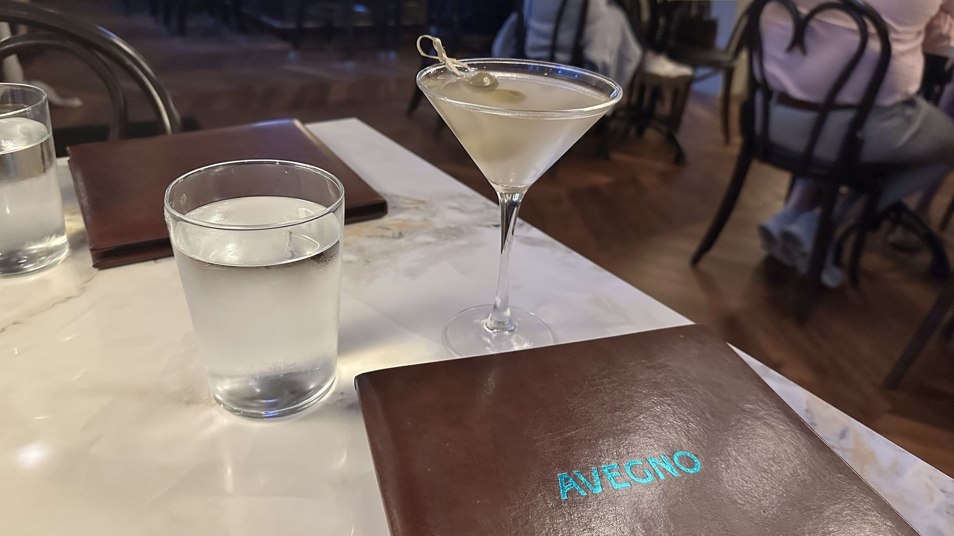 Martini glass with an olive sits on a white marble table beside a frosted glass of water; a brown menu rests nearby, with blurred restaurant patrons in the background.