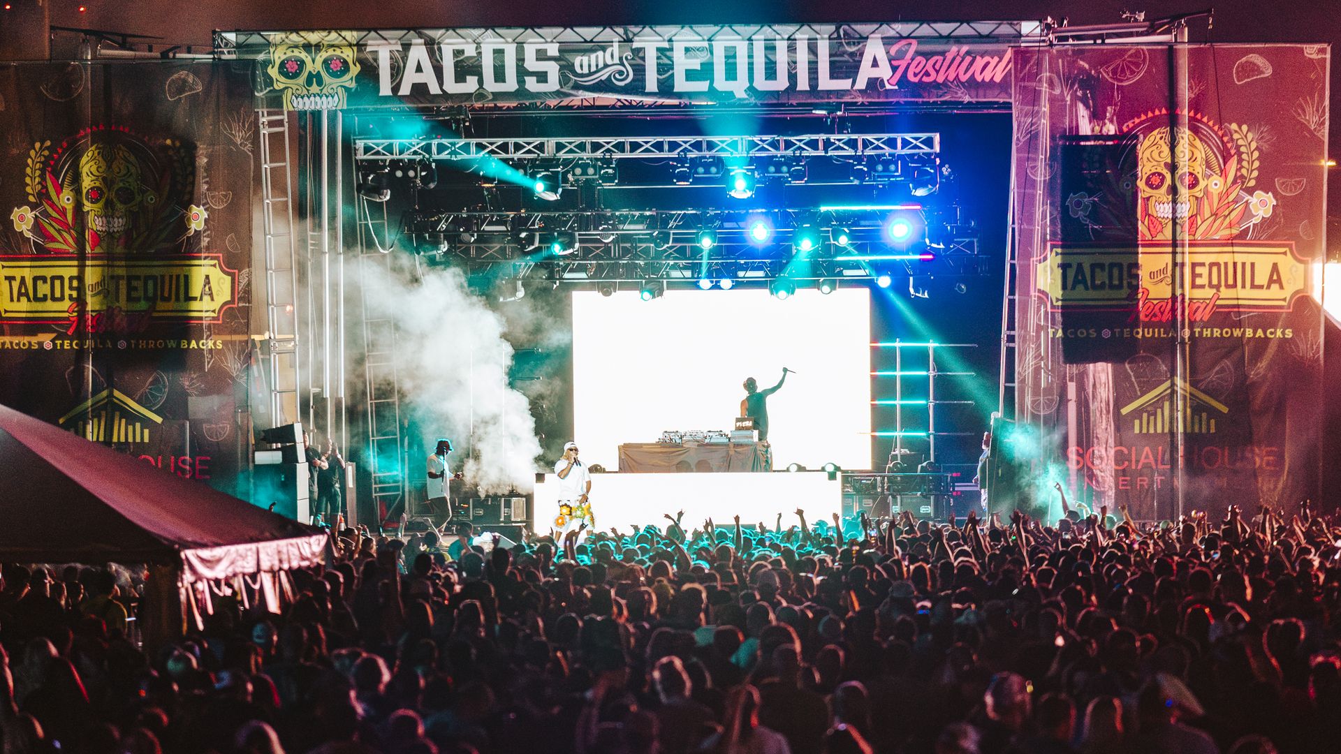 ChatGPT said: ChatGPT A large crowd watches a nighttime performance at the Tacos and Tequila Festival, with vibrant stage lighting, smoke effects, and performers silhouetted against a bright screen; banners with sugar skull artwork and the festival name frame the stage. 