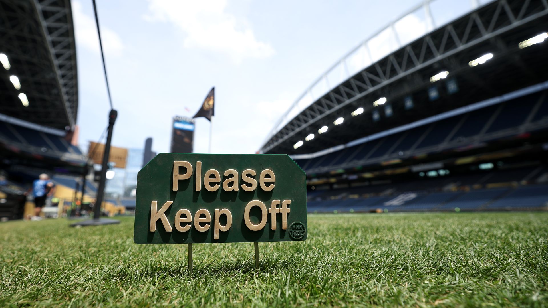 Closeup of grass and a "Please Keep Off" sign in the middle of a stadium with sky above. 