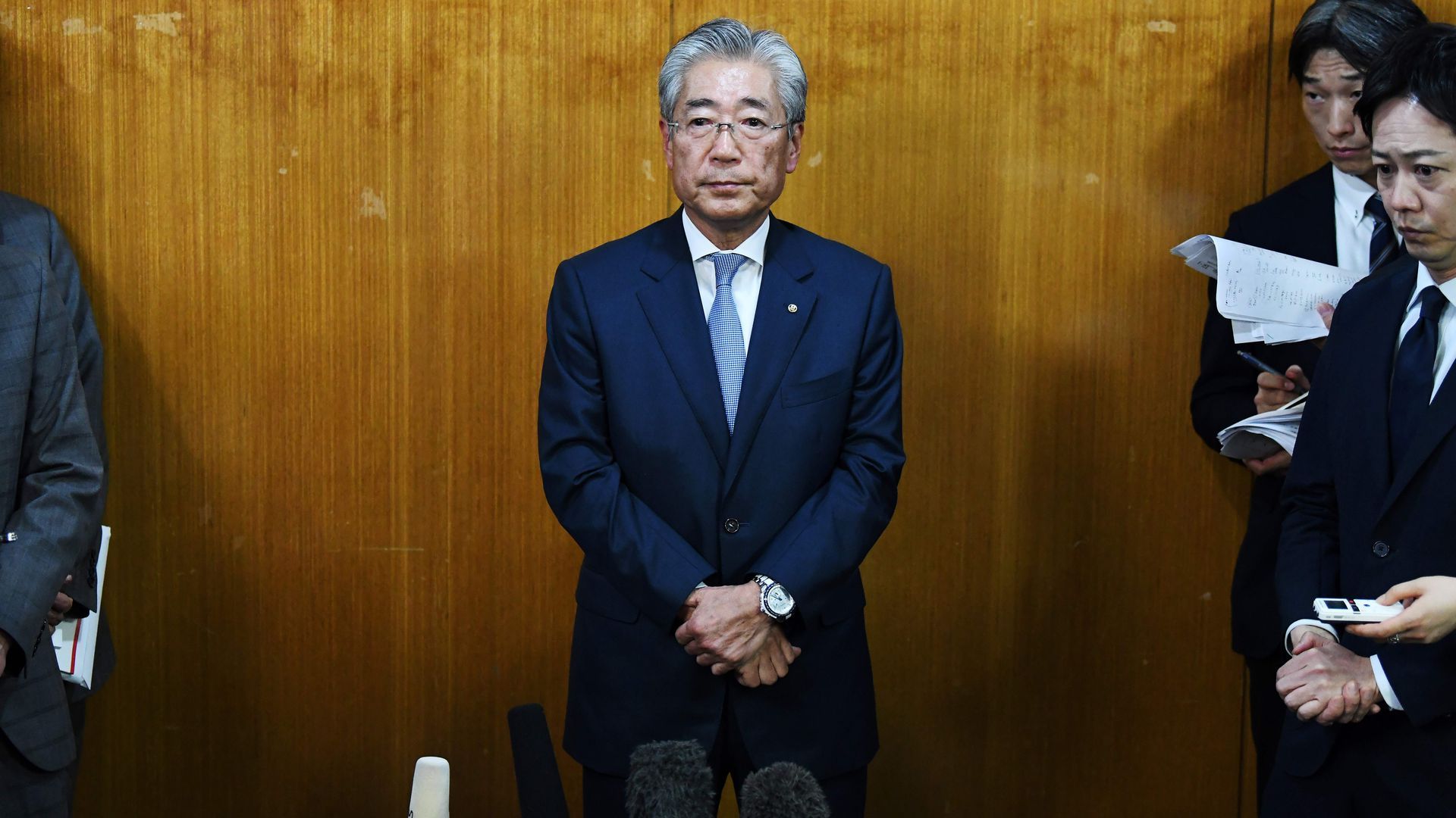 Tsunekazu Takeda stands in a blue suit with his arms folded and stands against a wooden wall. 