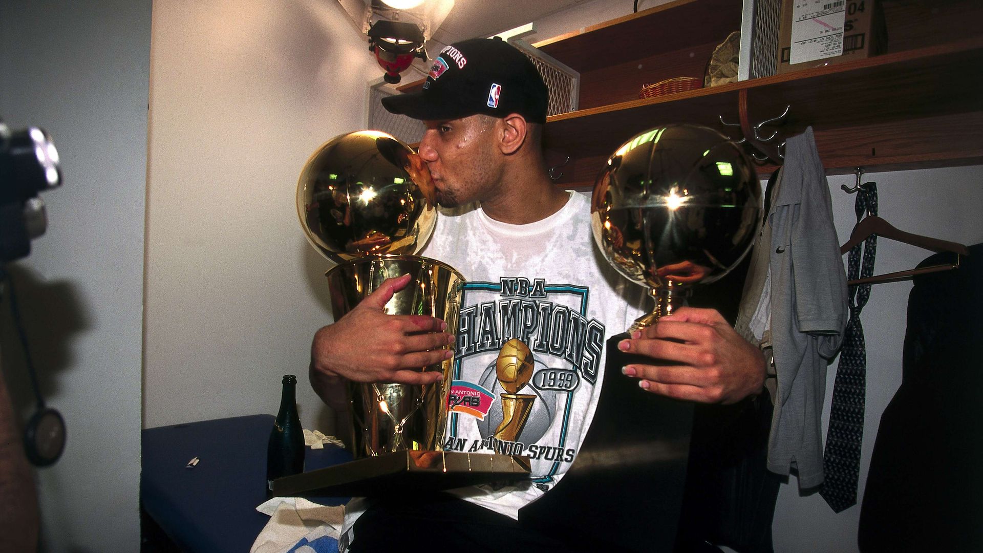 Man in a black NBA cap kisses two tall gold championship trophies in a locker room. He wears a white shirt displaying the title "NBA Champions 1999", with shelves, coats, and a bottle nearby.