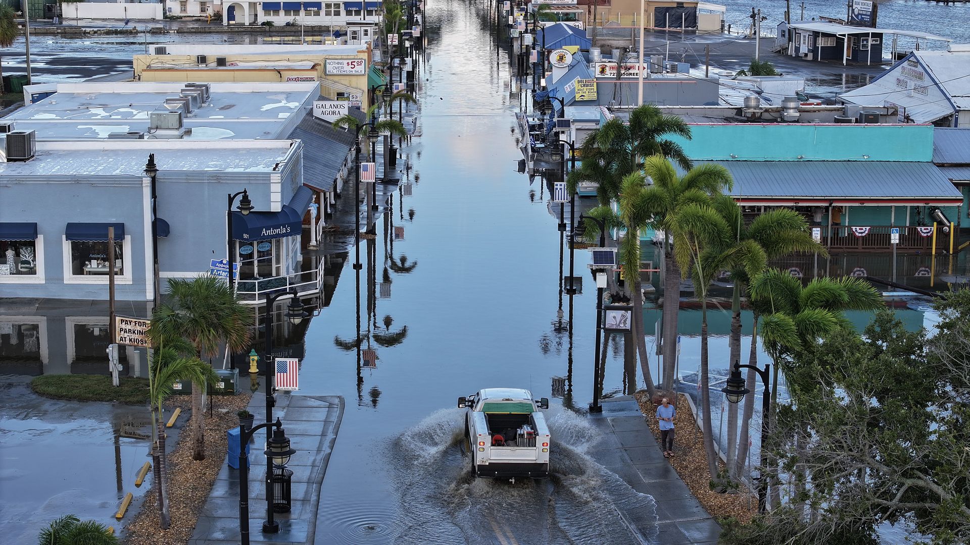 A white truck drives through a flooded road with businesses on each side.